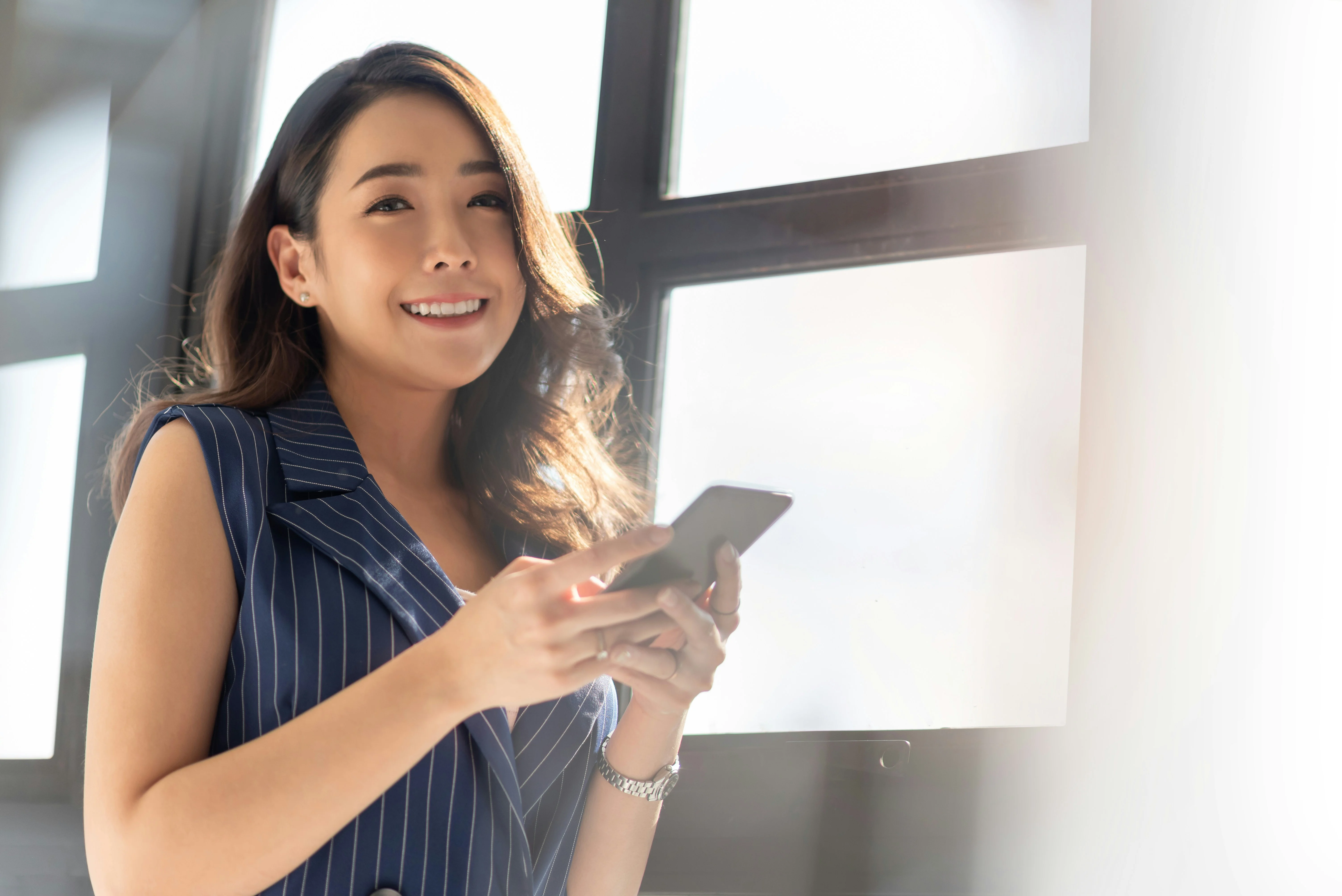 Smiling woman in a pinstripe outfit holds a smartphone, standing near large windows with bright natural light.