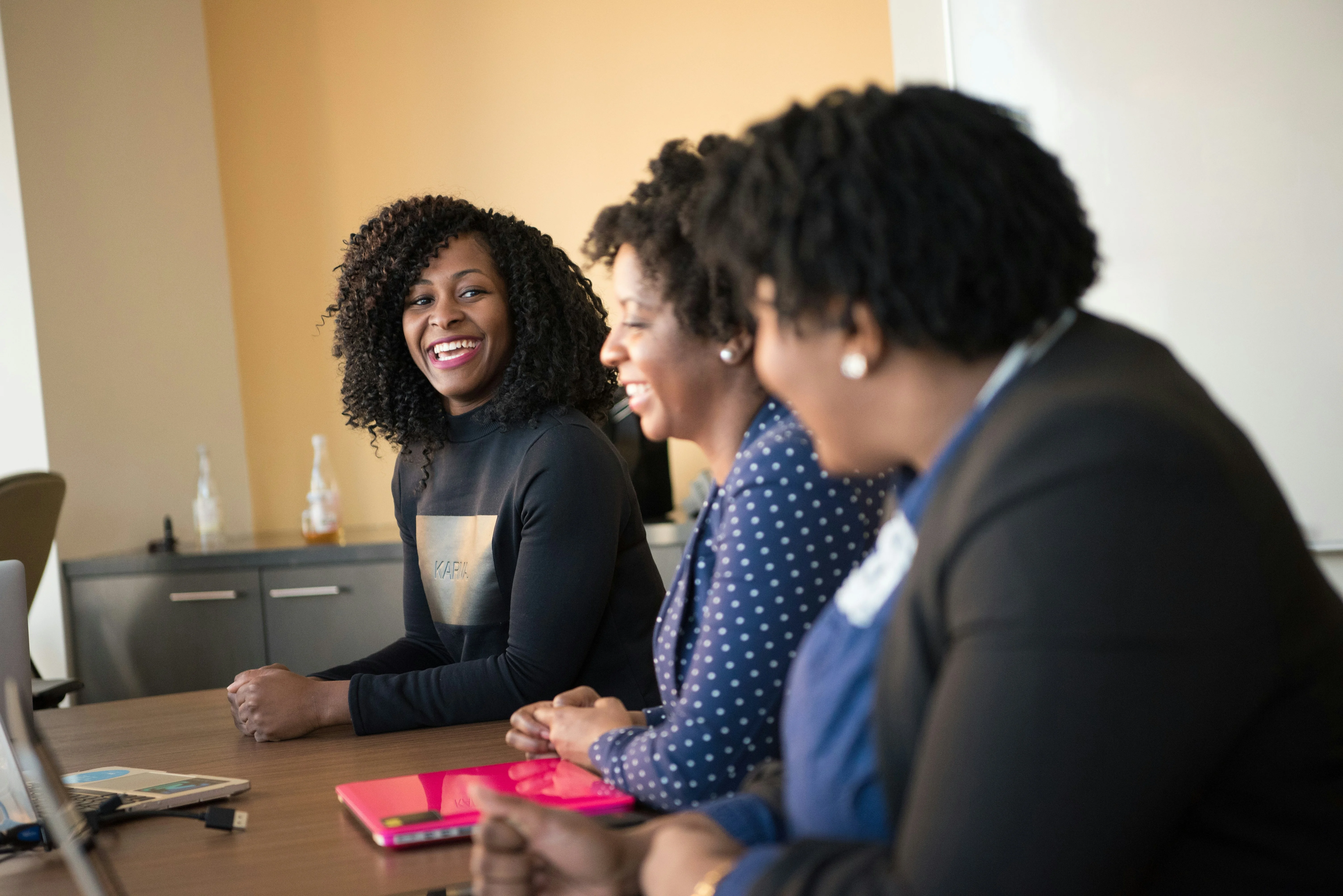 Three women sitting at a conference table, smiling and engaged in conversation, with notebooks and a laptop in front of them.