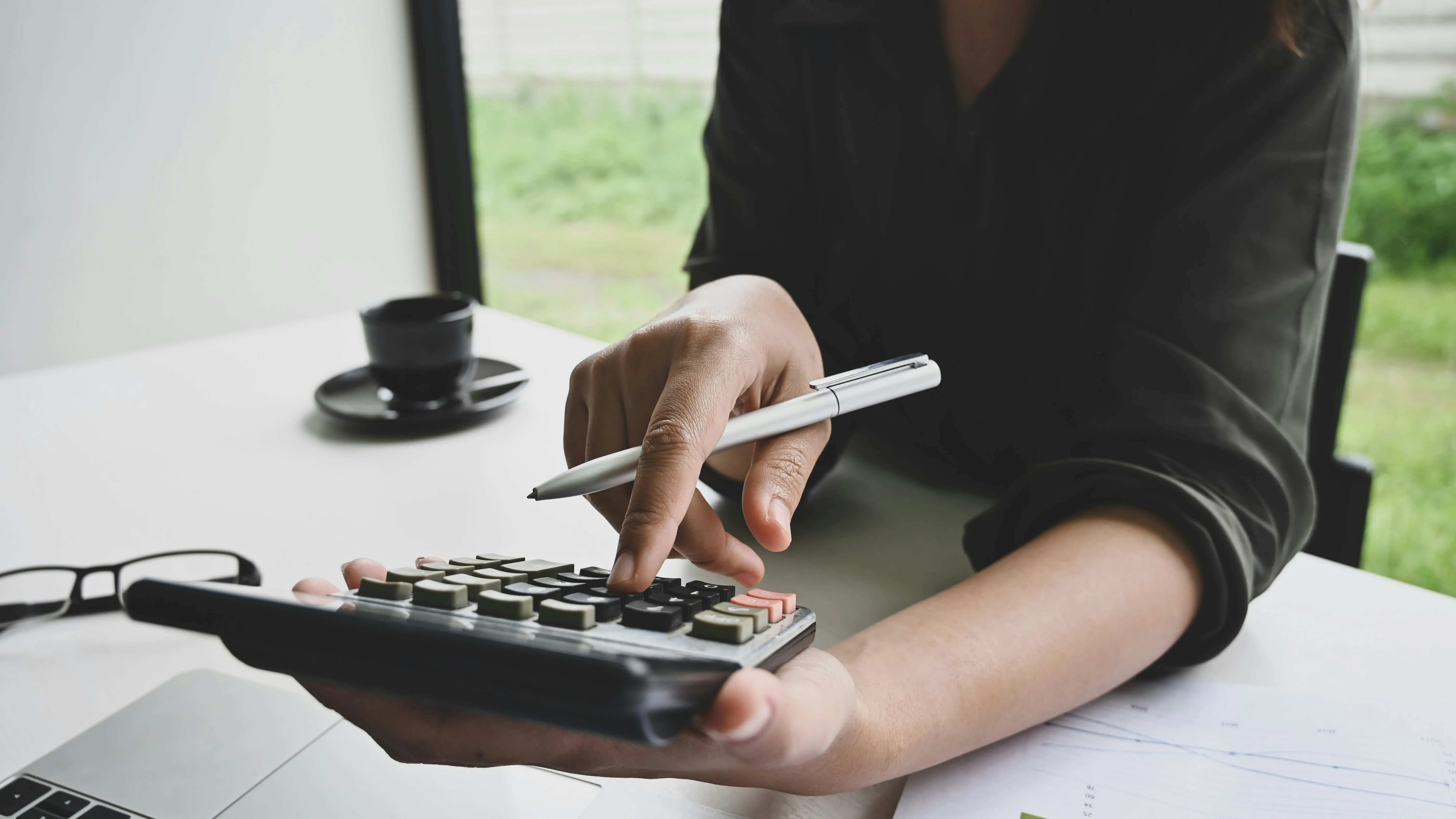 Person using a calculator at a desk, holding a pen. A coffee cup, glasses, and documents are nearby.