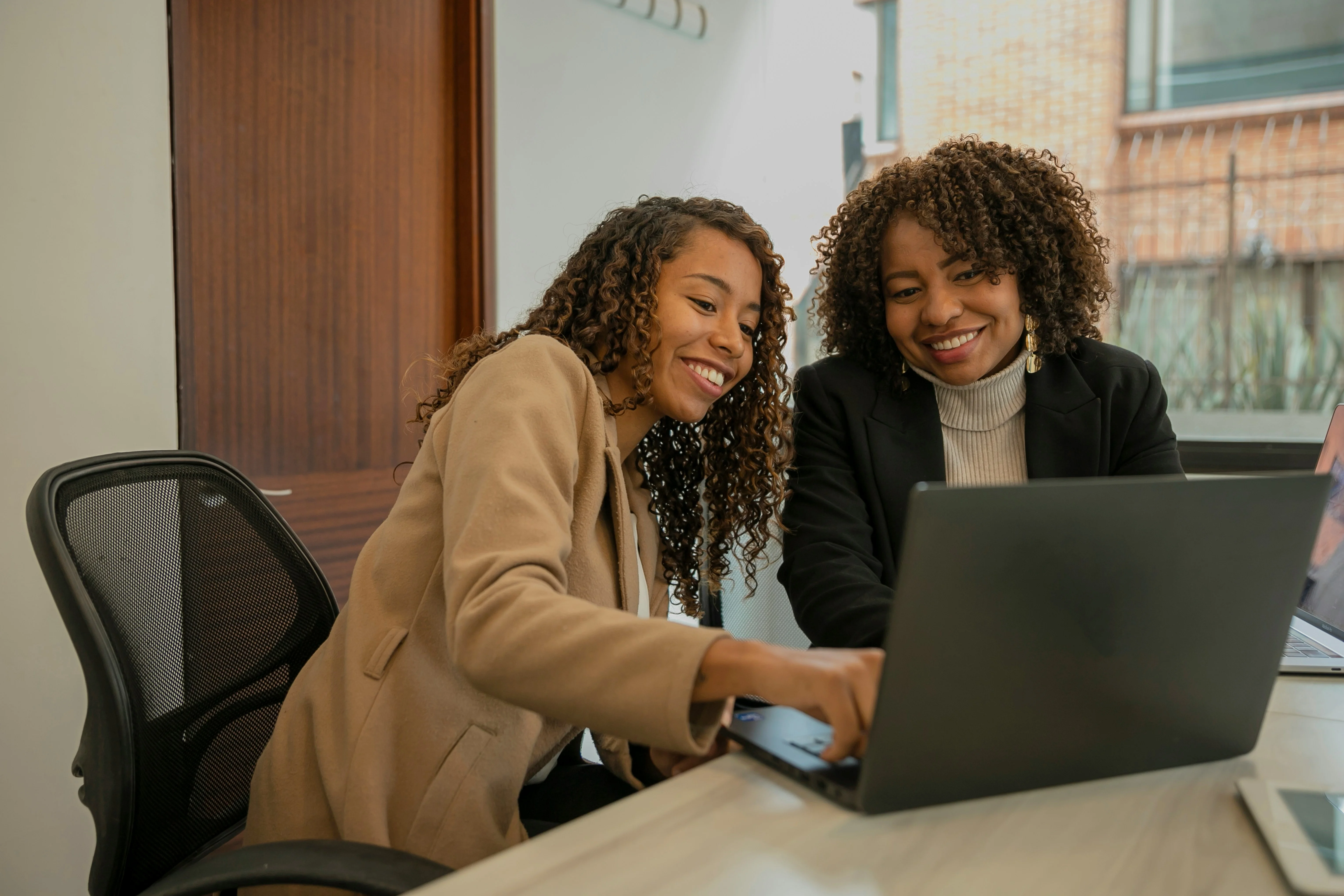 Two women with curly hair smiling and working together on a laptop at a desk in an office setting.