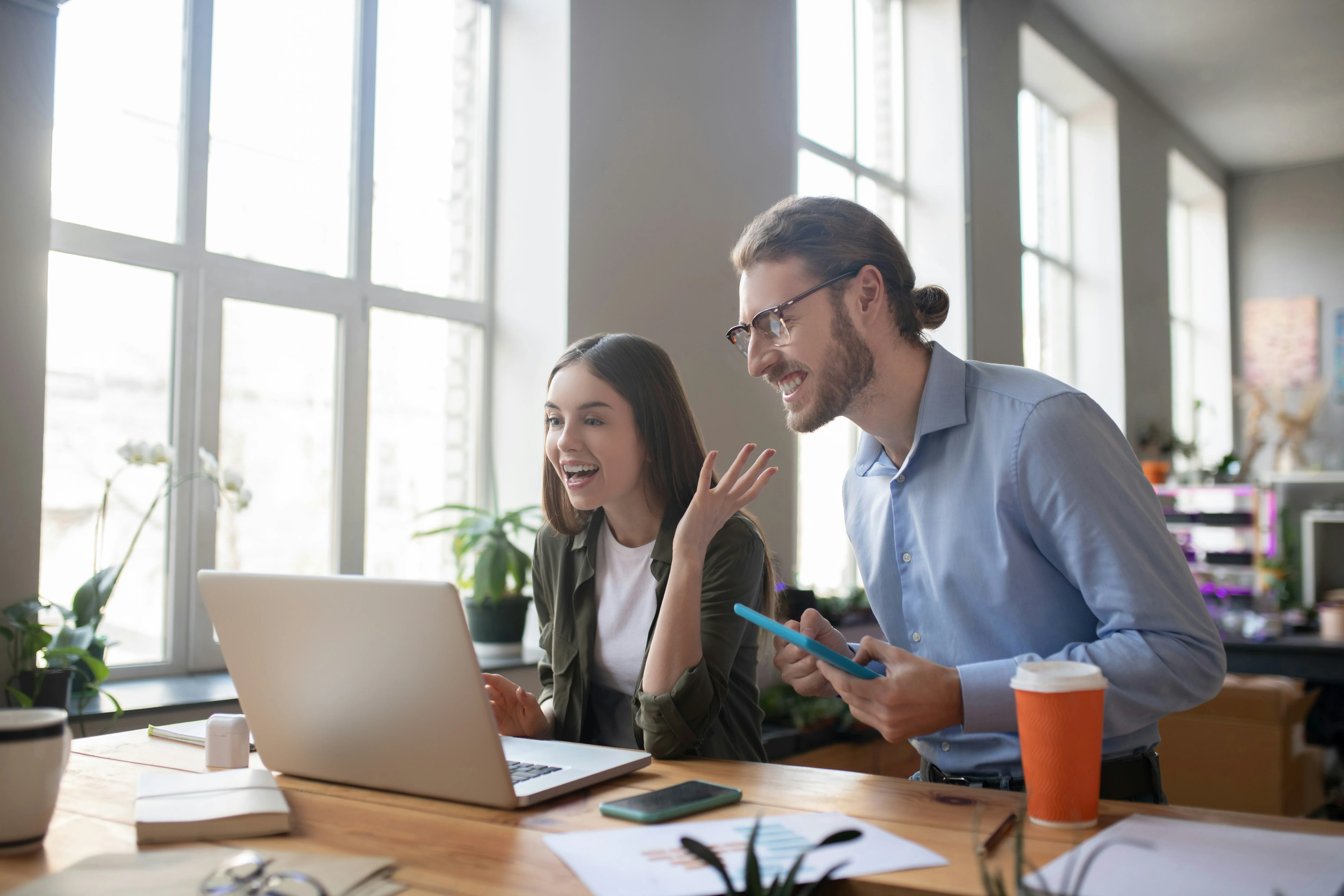 Two people smiling and video chatting on a laptop in a bright office with large windows. A coffee cup and potted plants are on the desk.