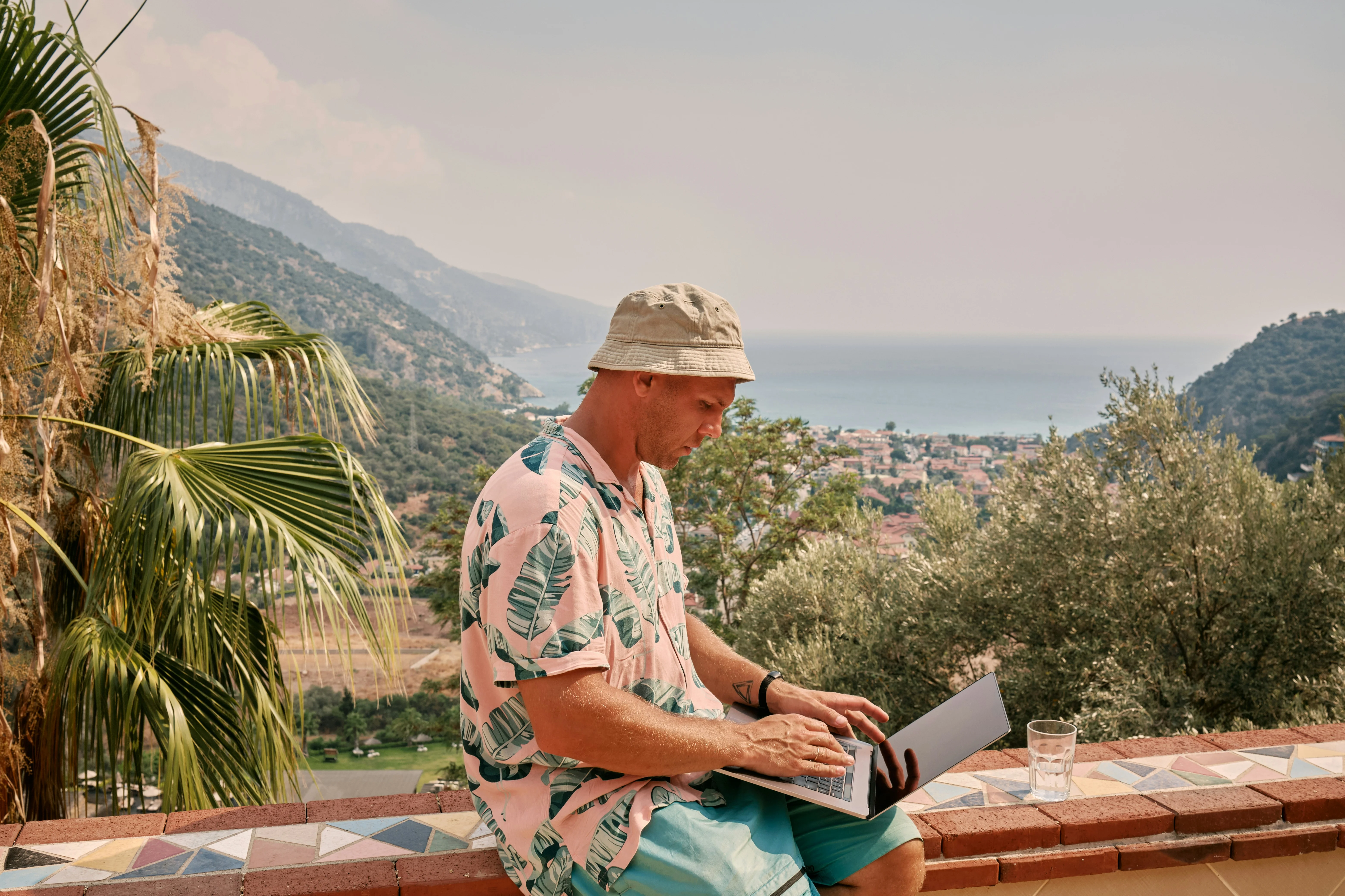 Man in tropical shirt and bucket hat using a laptop on a terrace with a scenic view of mountains and the sea.
