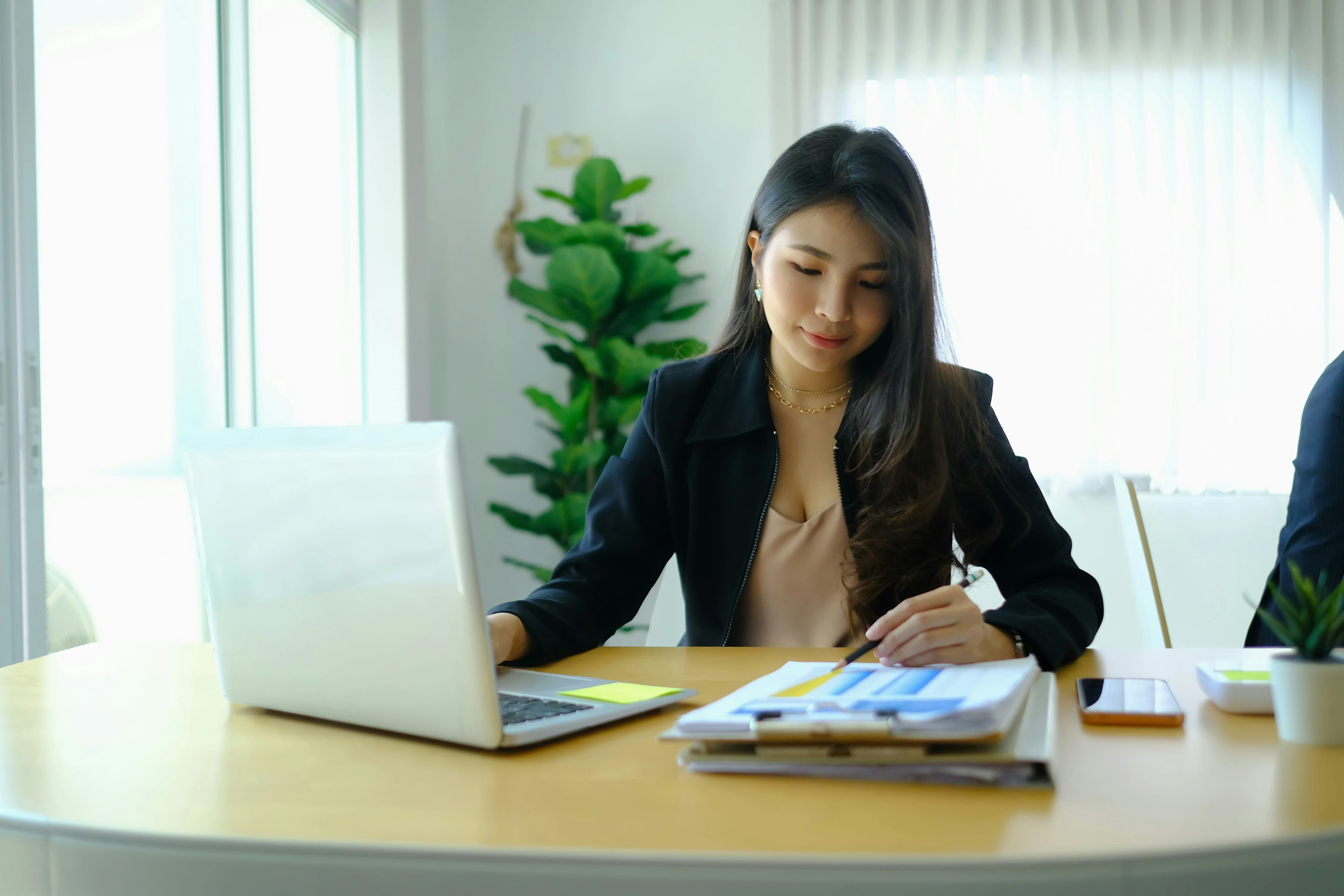 Woman in a black blazer working at a desk with a laptop and documents, in a bright office with a plant in the background.