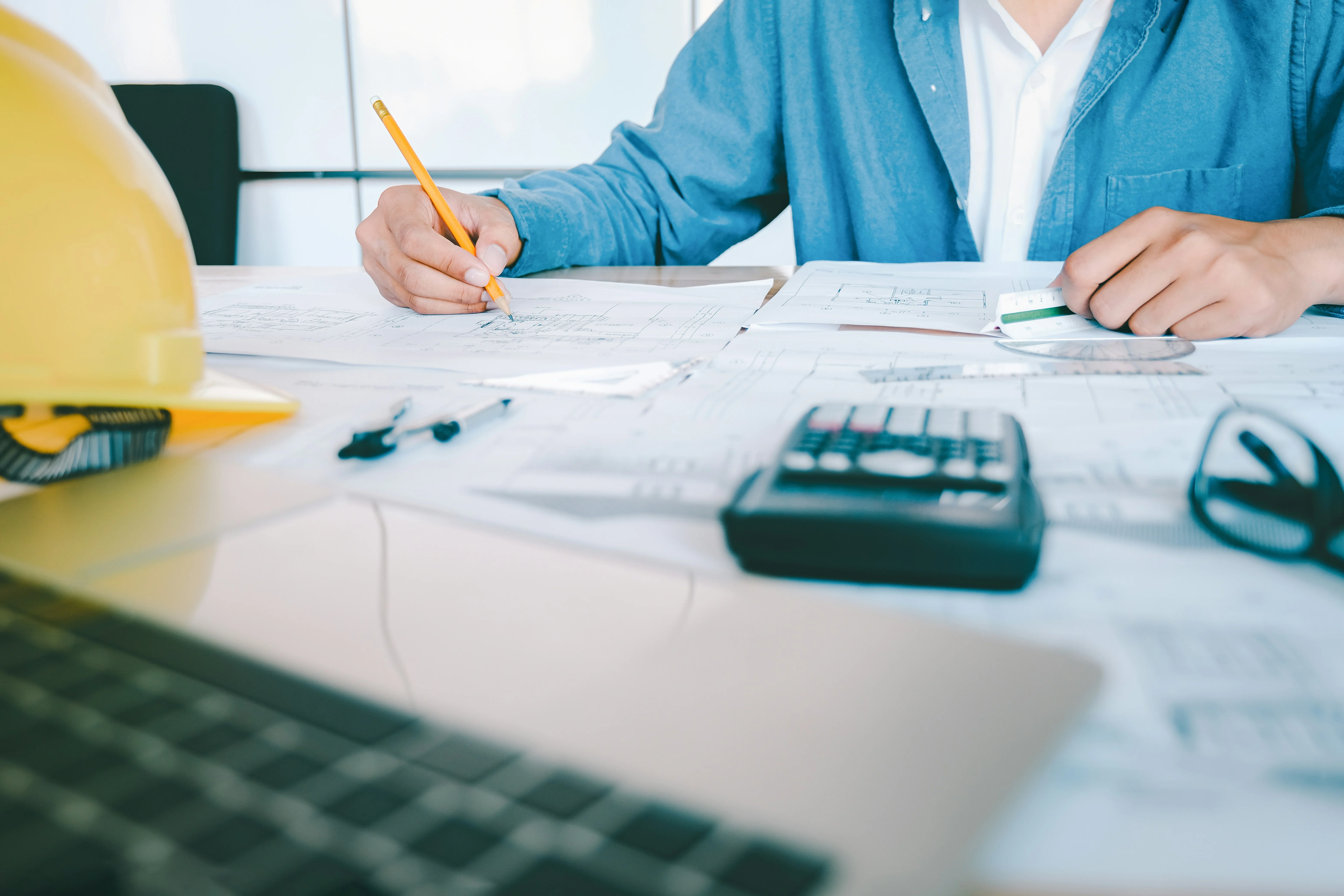 Person in a blue shirt working on architectural plans at a desk with a calculator, helmet, and glasses nearby.
