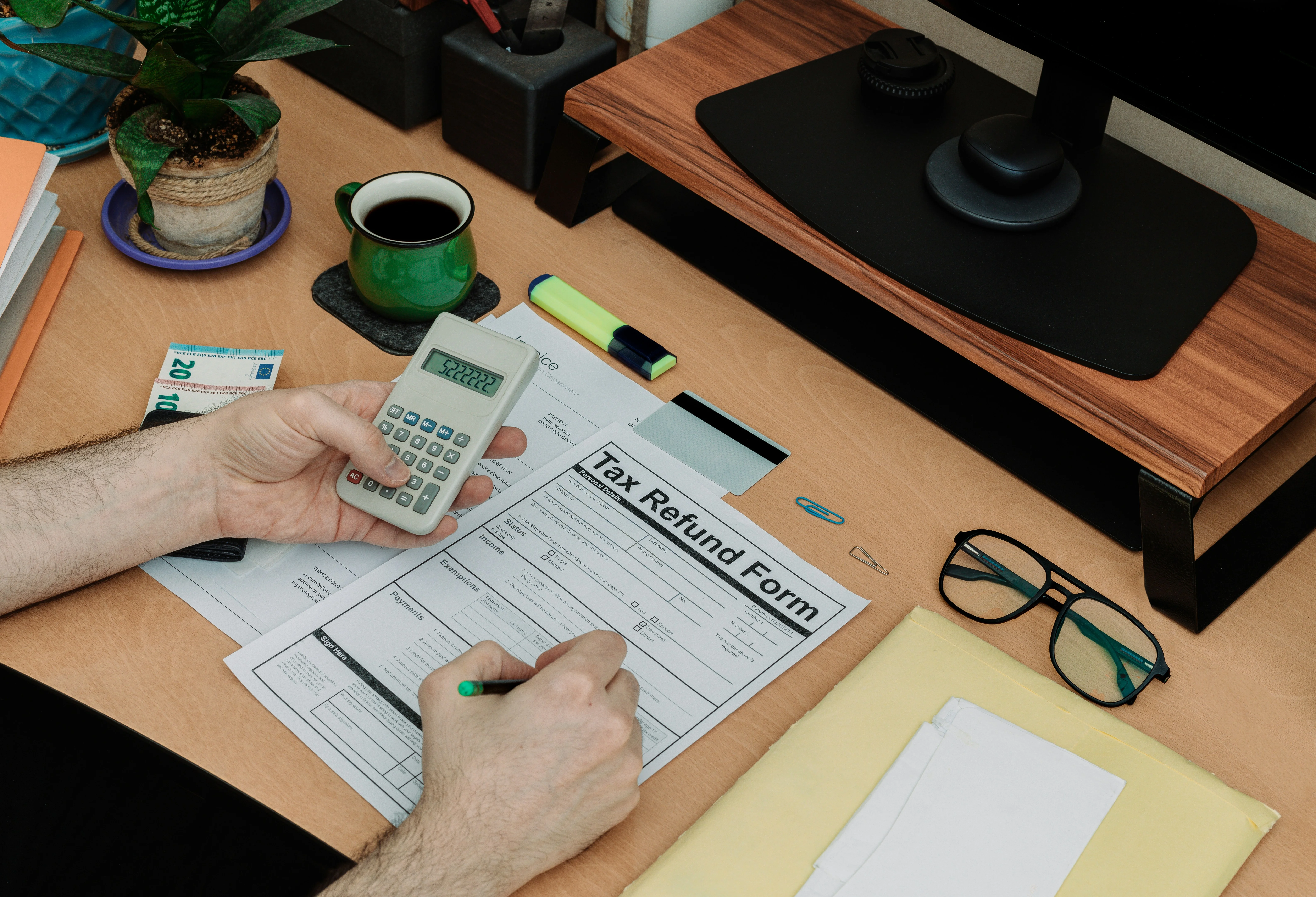 Person filling out a tax refund form at a desk with a calculator, coffee, glasses, and stationery.