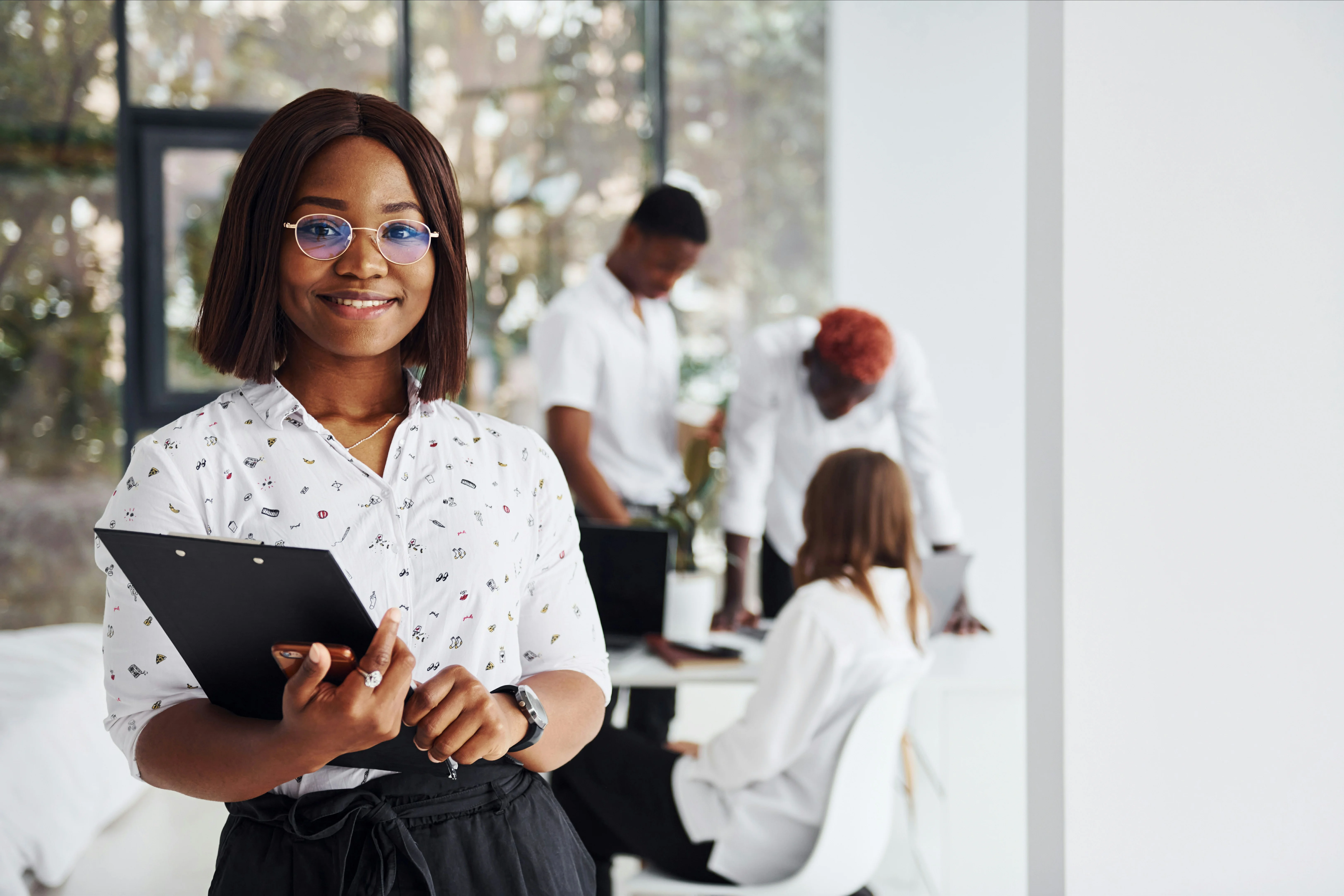 Woman smiling and holding a clipboard in an office, with three colleagues collaborating in the background.