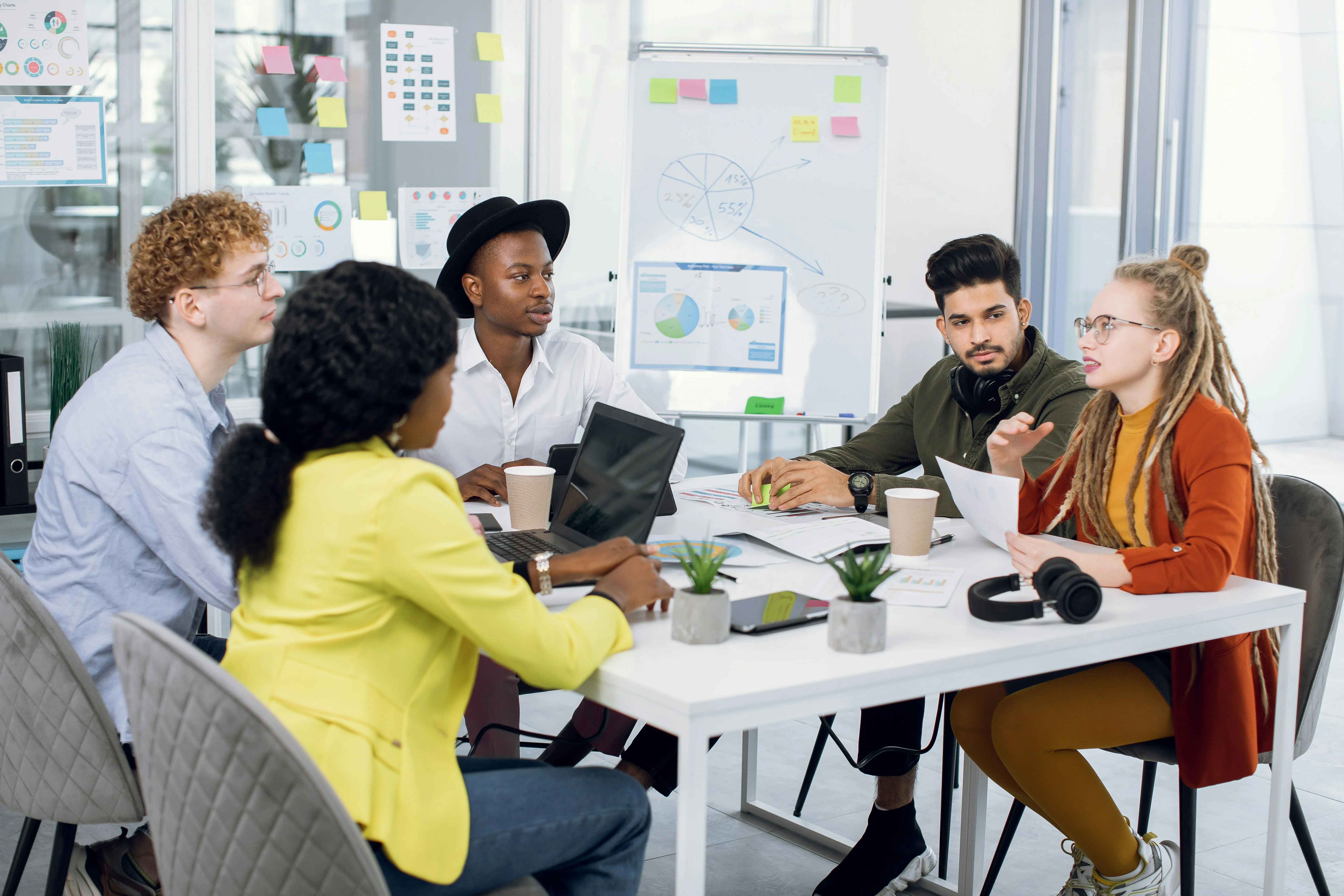 A diverse group of people engaged in a meeting around a table with laptops, charts, and a whiteboard in a modern office setting.
