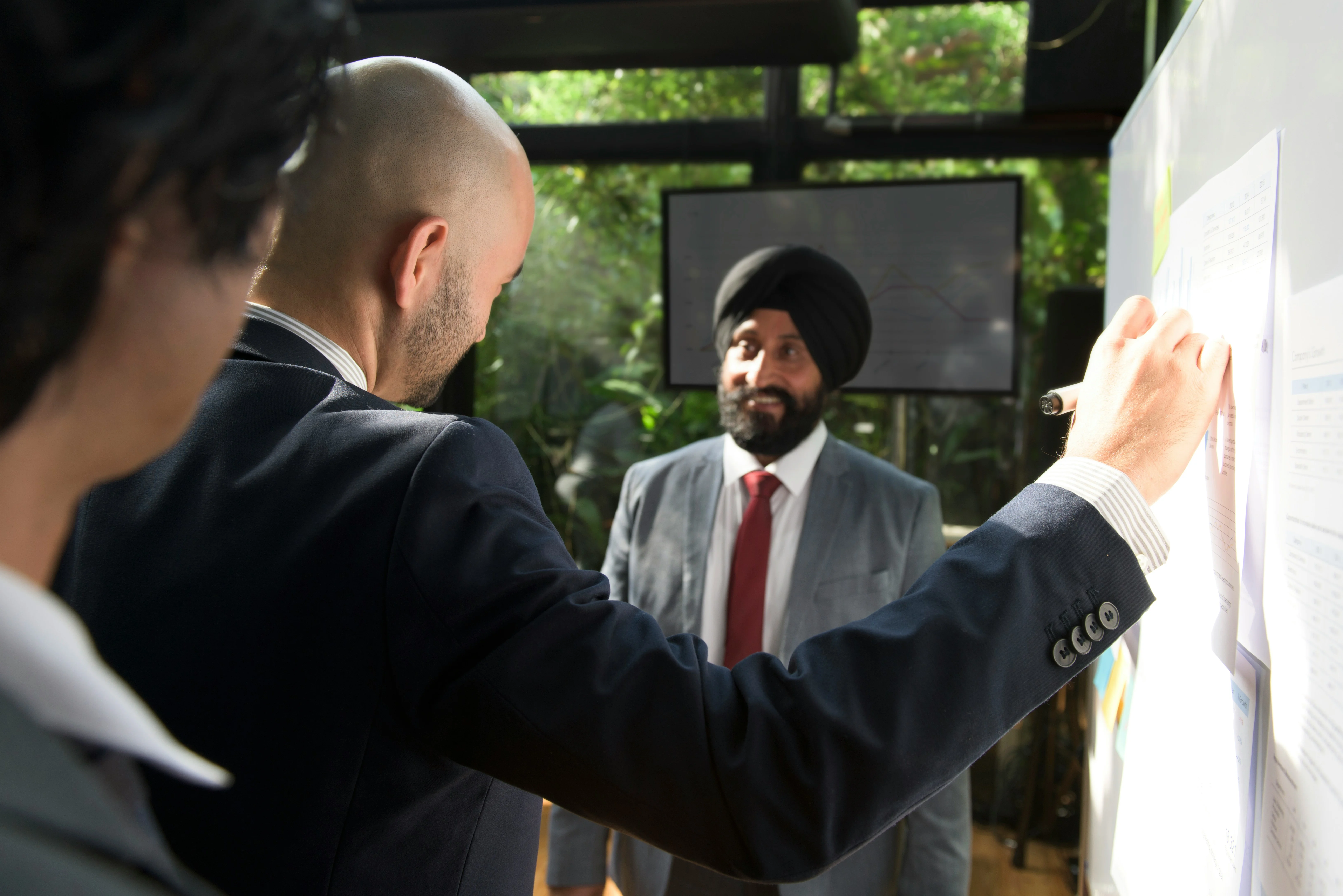 Three professionals in a meeting room, one writing on a whiteboard while another observes, with a screen and greenery in the background.