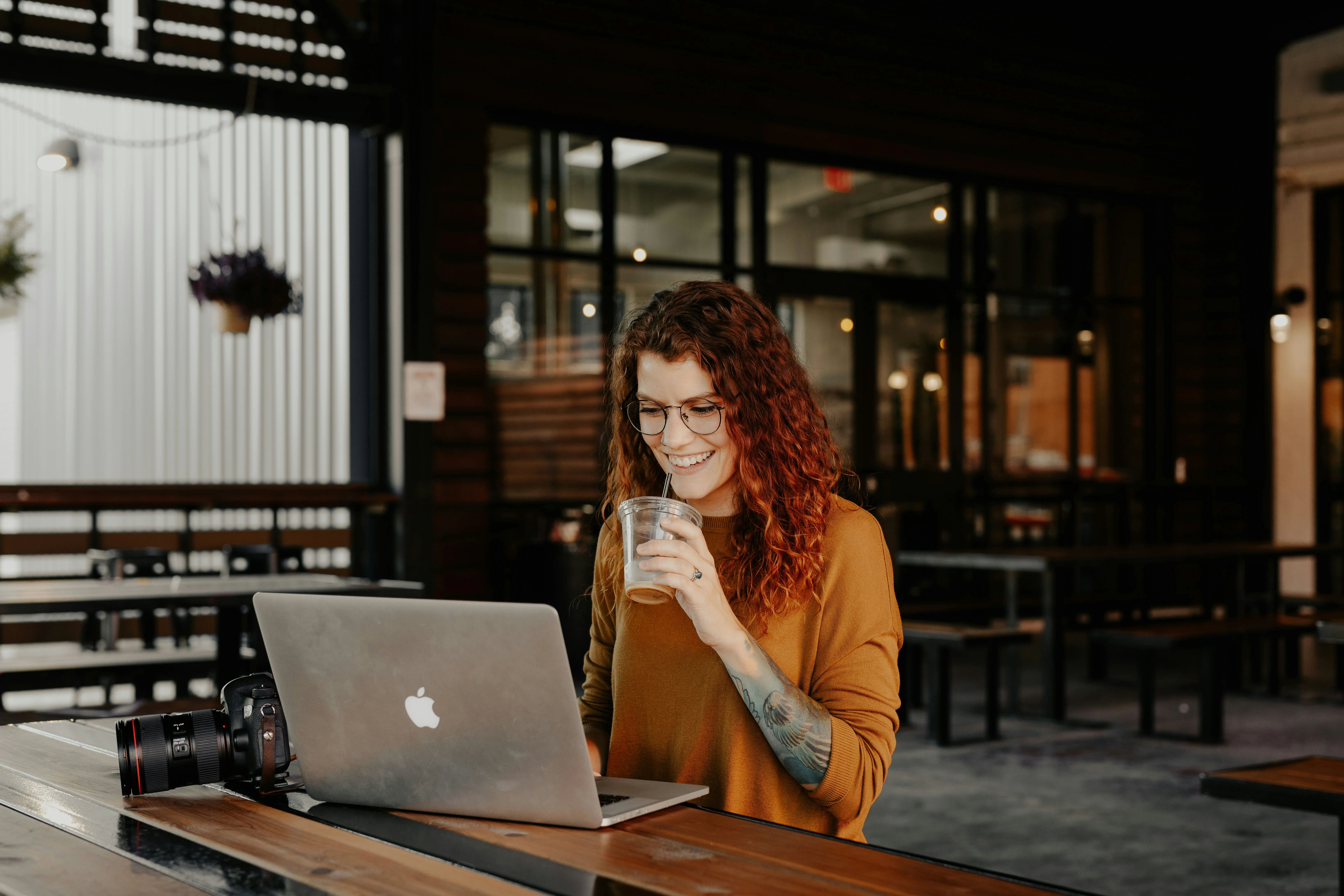 A woman with curly hair and glasses, wearing a mustard sweater, sits at a table with a laptop and camera, sipping a drink in a modern café.
