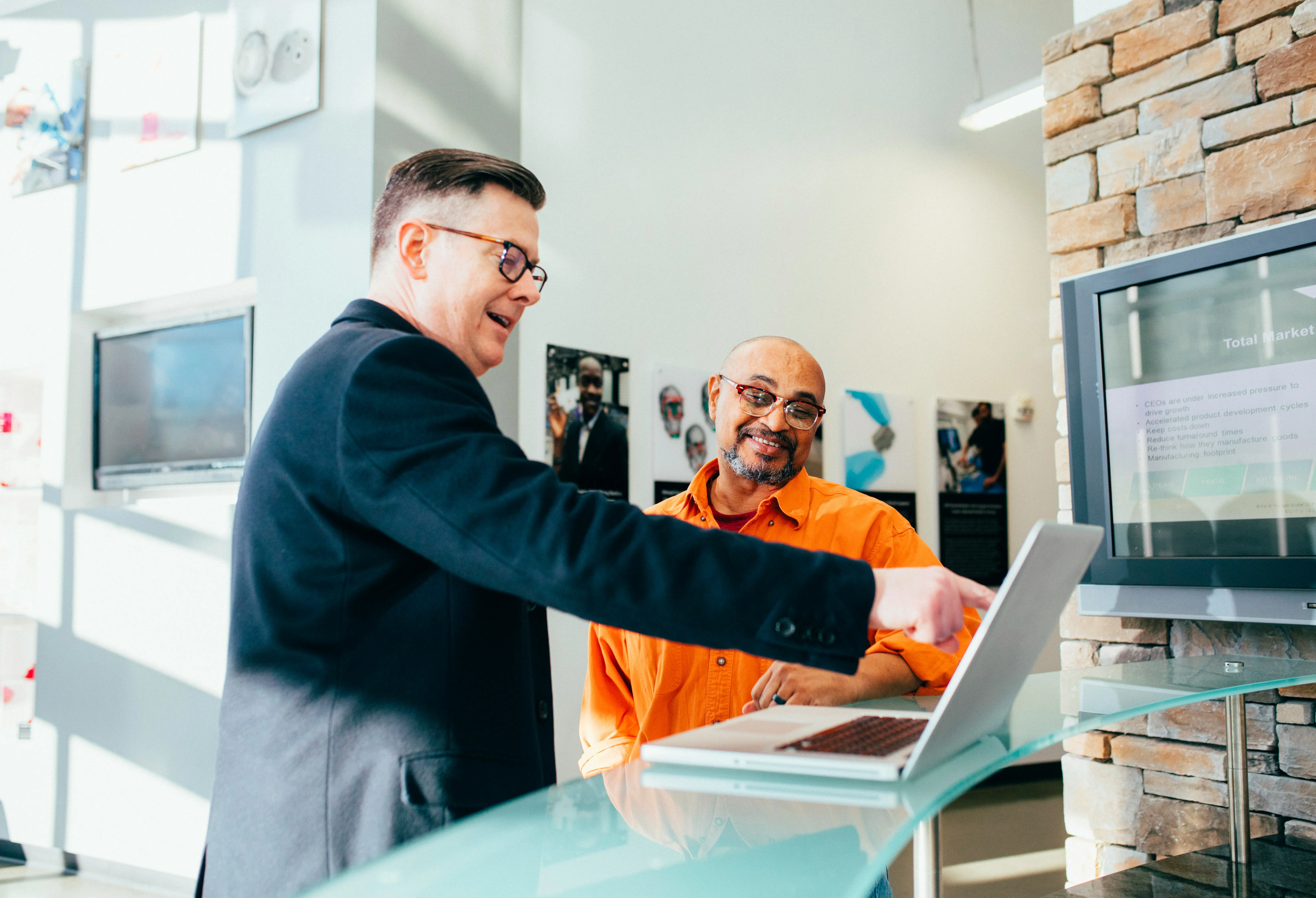 Two men in a modern office setting, one in a suit pointing at a laptop screen, while the other, in an orange shirt, smiles.