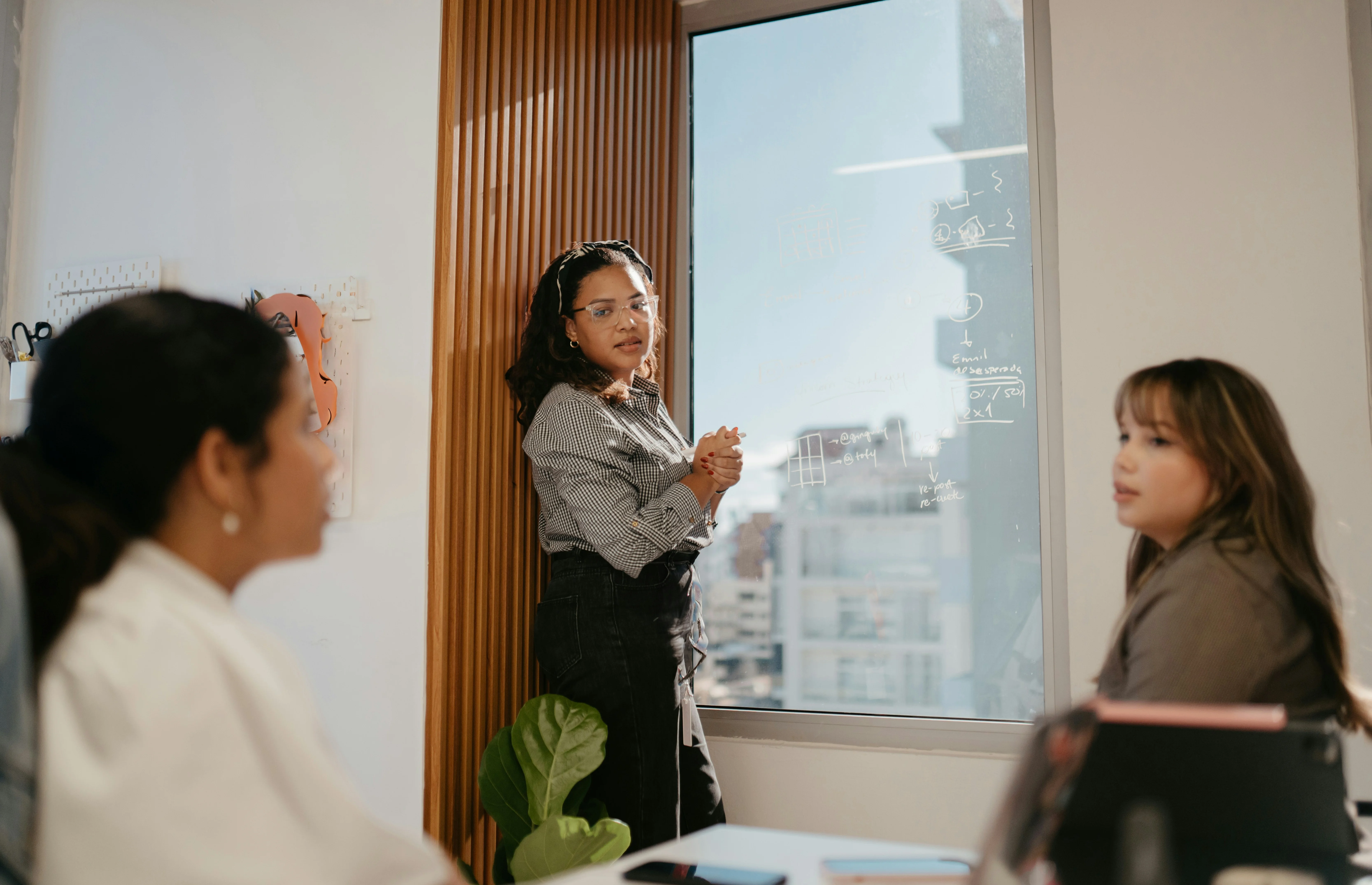 A woman stands explaining near a window with notes on it, while two people are seated, listening attentively in a well-lit room.