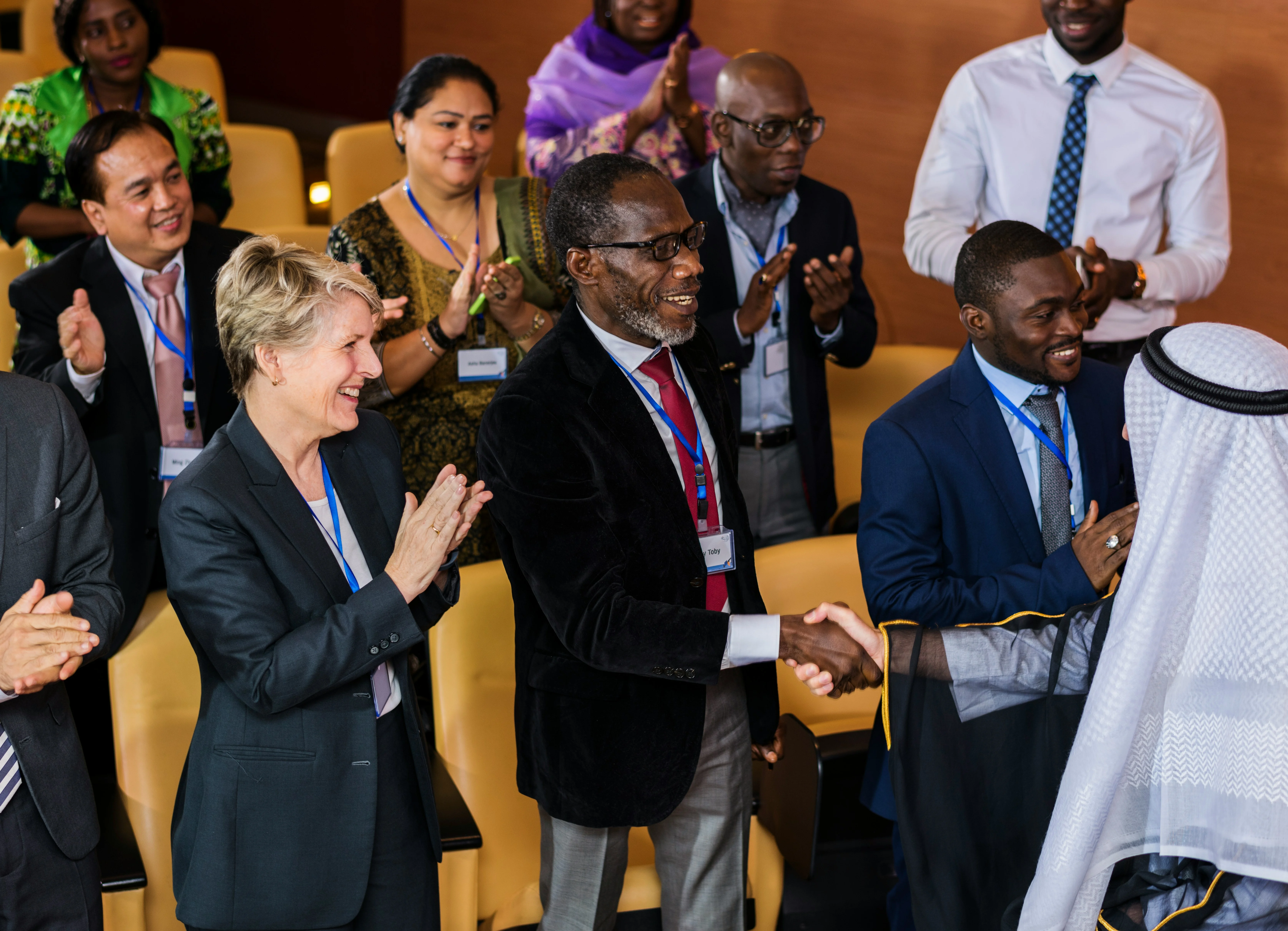 Diverse group of people in formal attire clapping, with two men shaking hands in the foreground at an event.