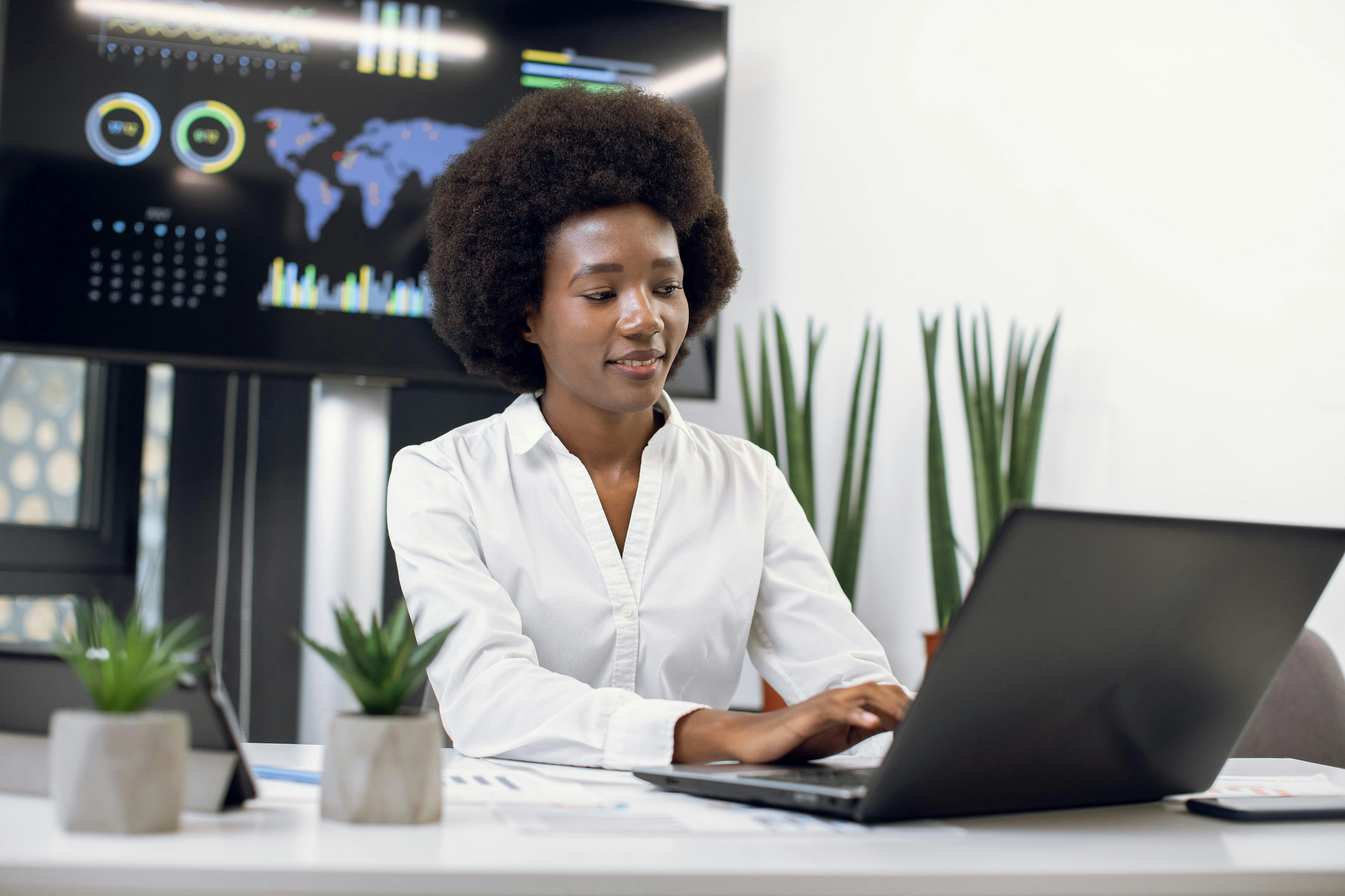 A woman in a white shirt works on a laptop at a desk with potted plants. A screen displays data and a world map in the background.