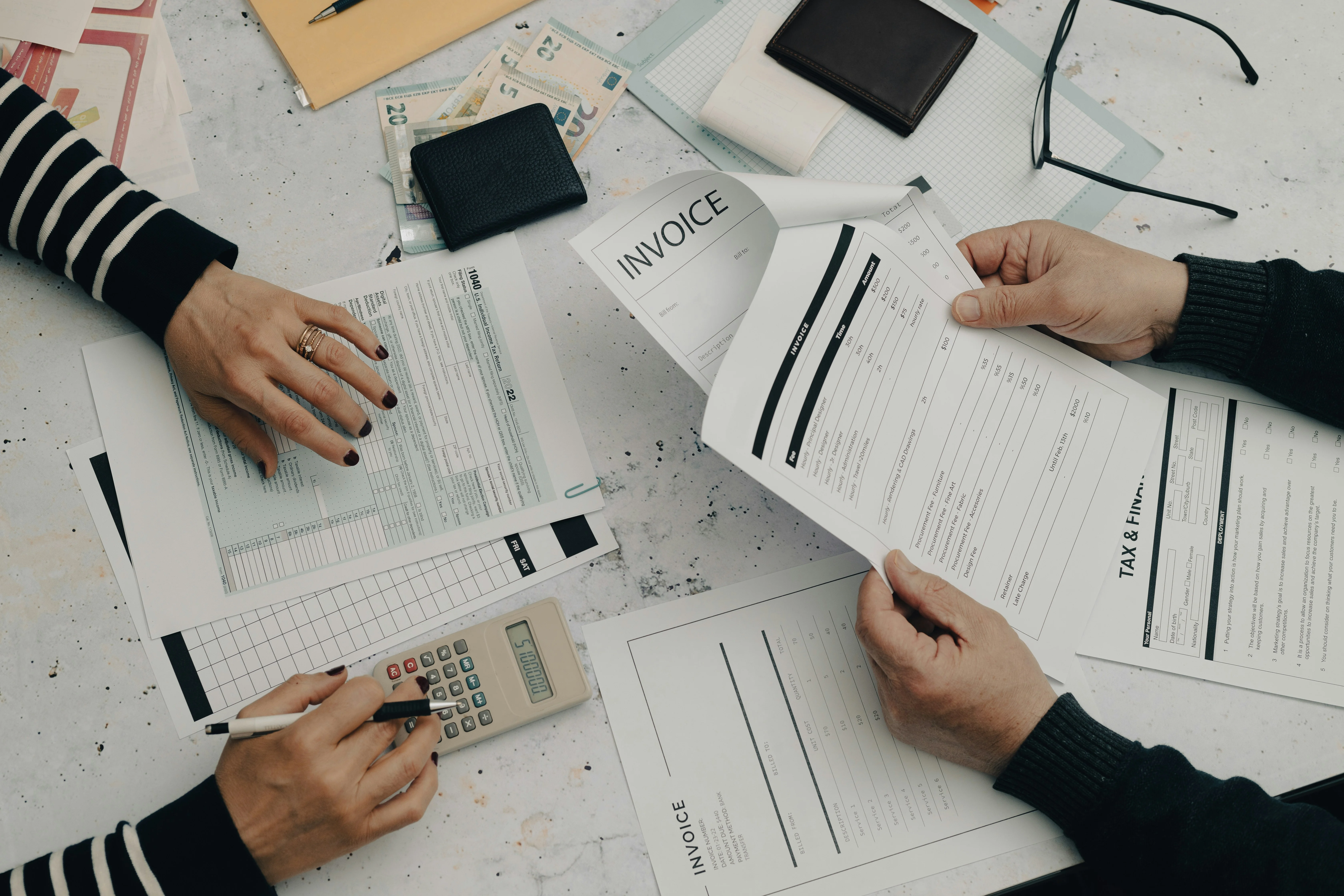 Two people working at a desk with invoices, tax forms, a calculator, and stationery, focusing on financial paperwork.