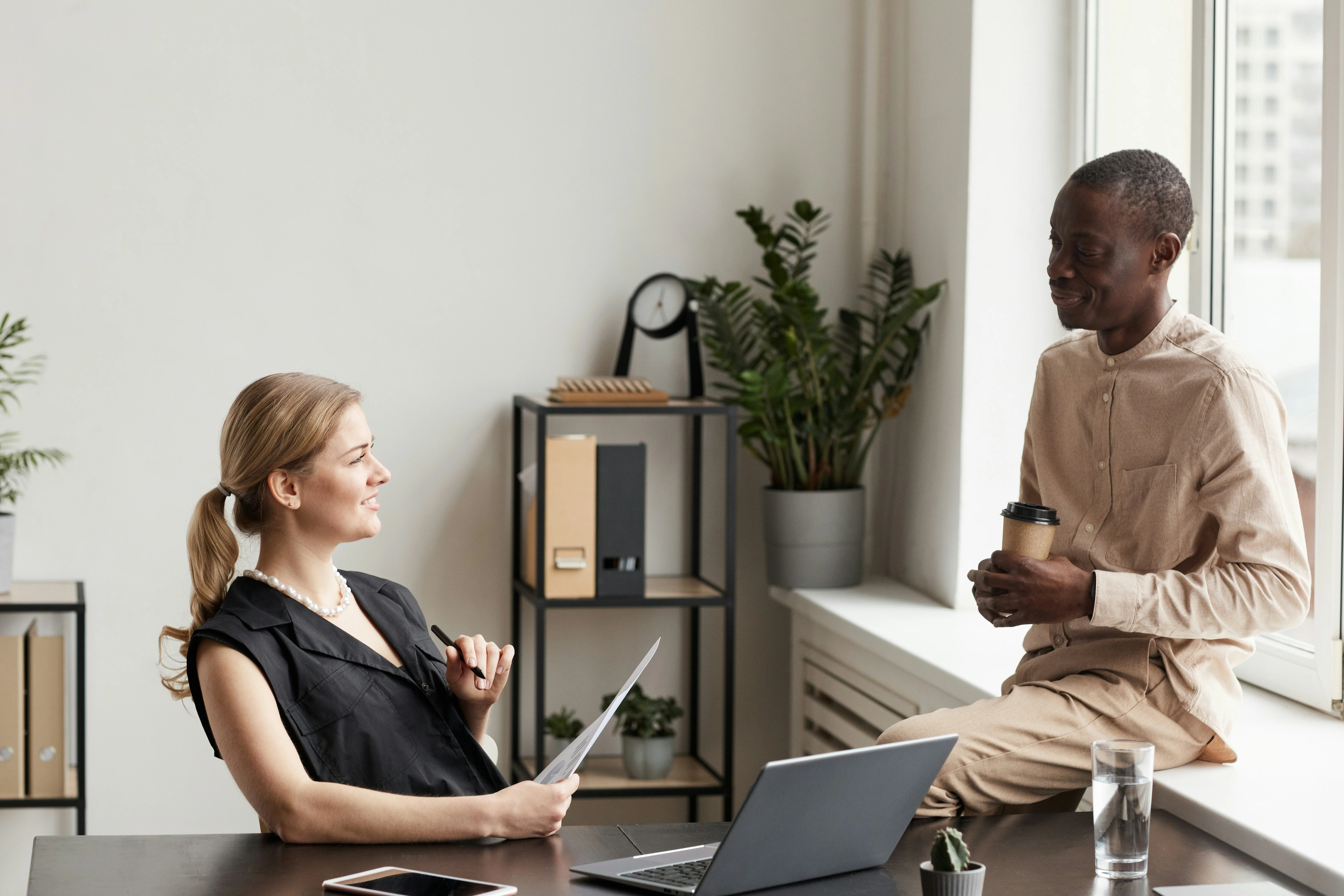 Two people in an office, one seated at a desk with papers, the other sitting on a windowsill holding a coffee cup.