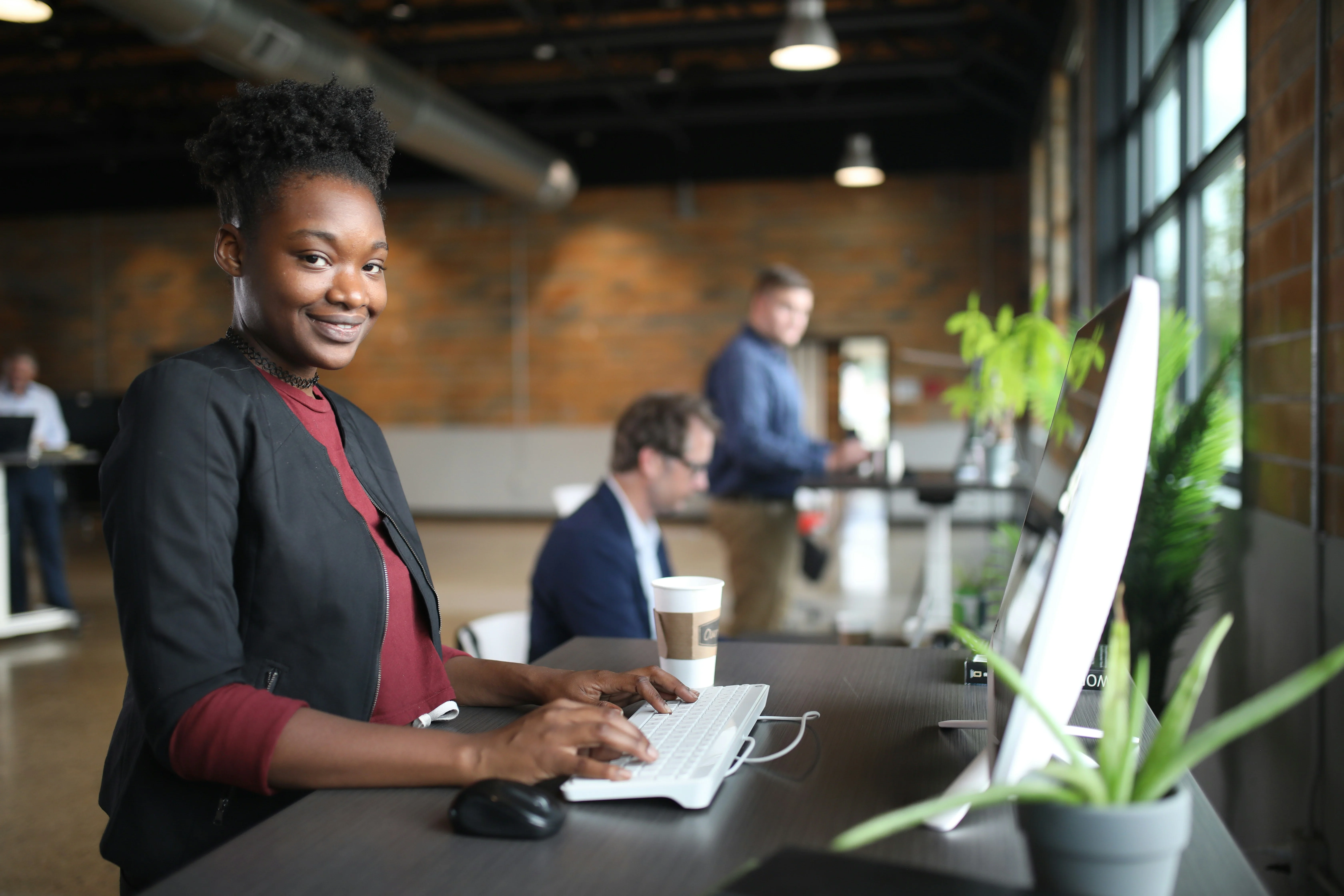 Woman smiling at desk with computer, holding a keyboard. Two colleagues work in the background. Office setting with natural lighting and plants.