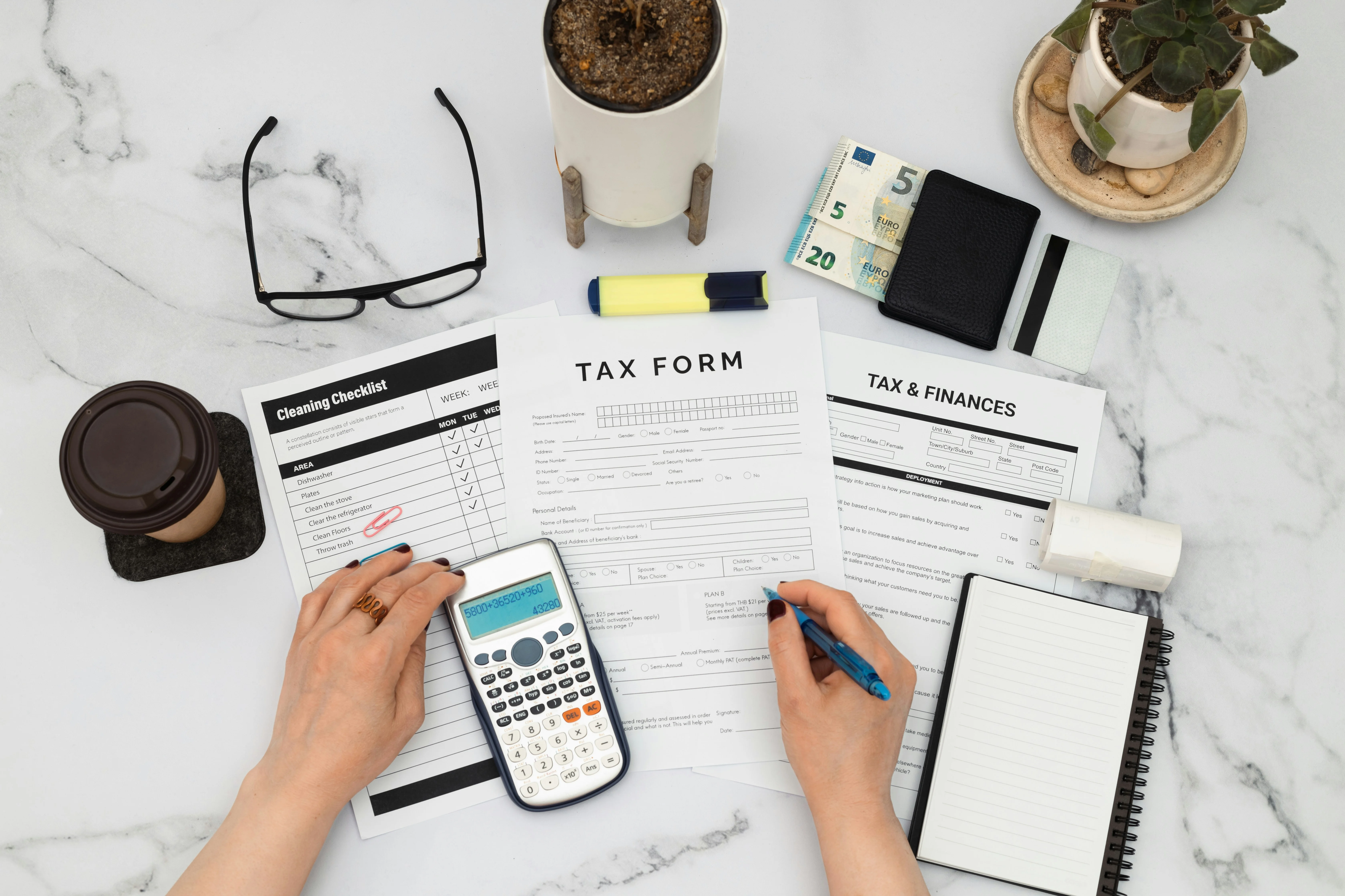 Person filling out tax forms on a desk with a calculator, glasses, coffee cup, wallet with cash, and potted plants.