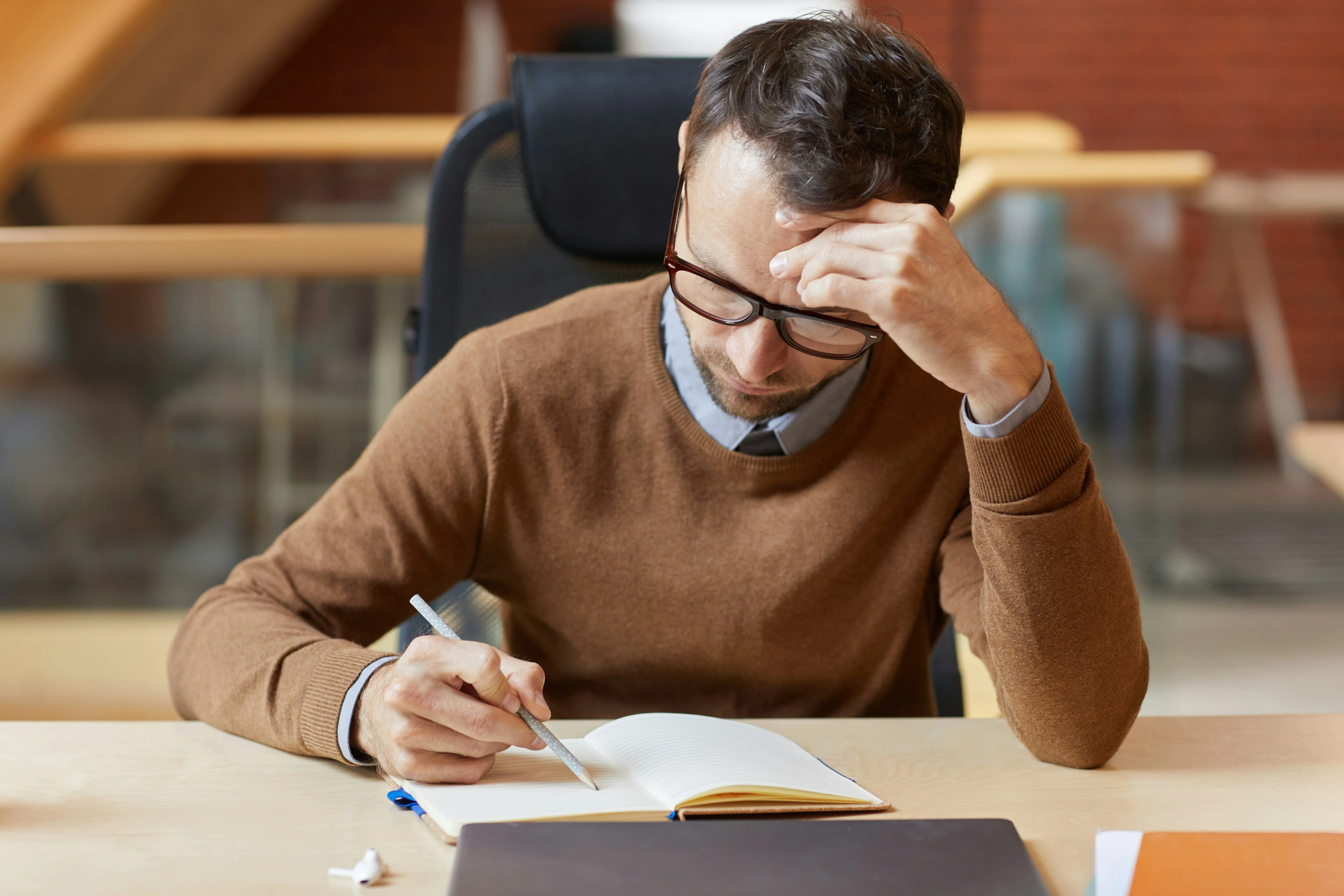 Man wearing glasses sits at a desk, writing in a notebook with a thoughtful expression, resting his head on one hand.