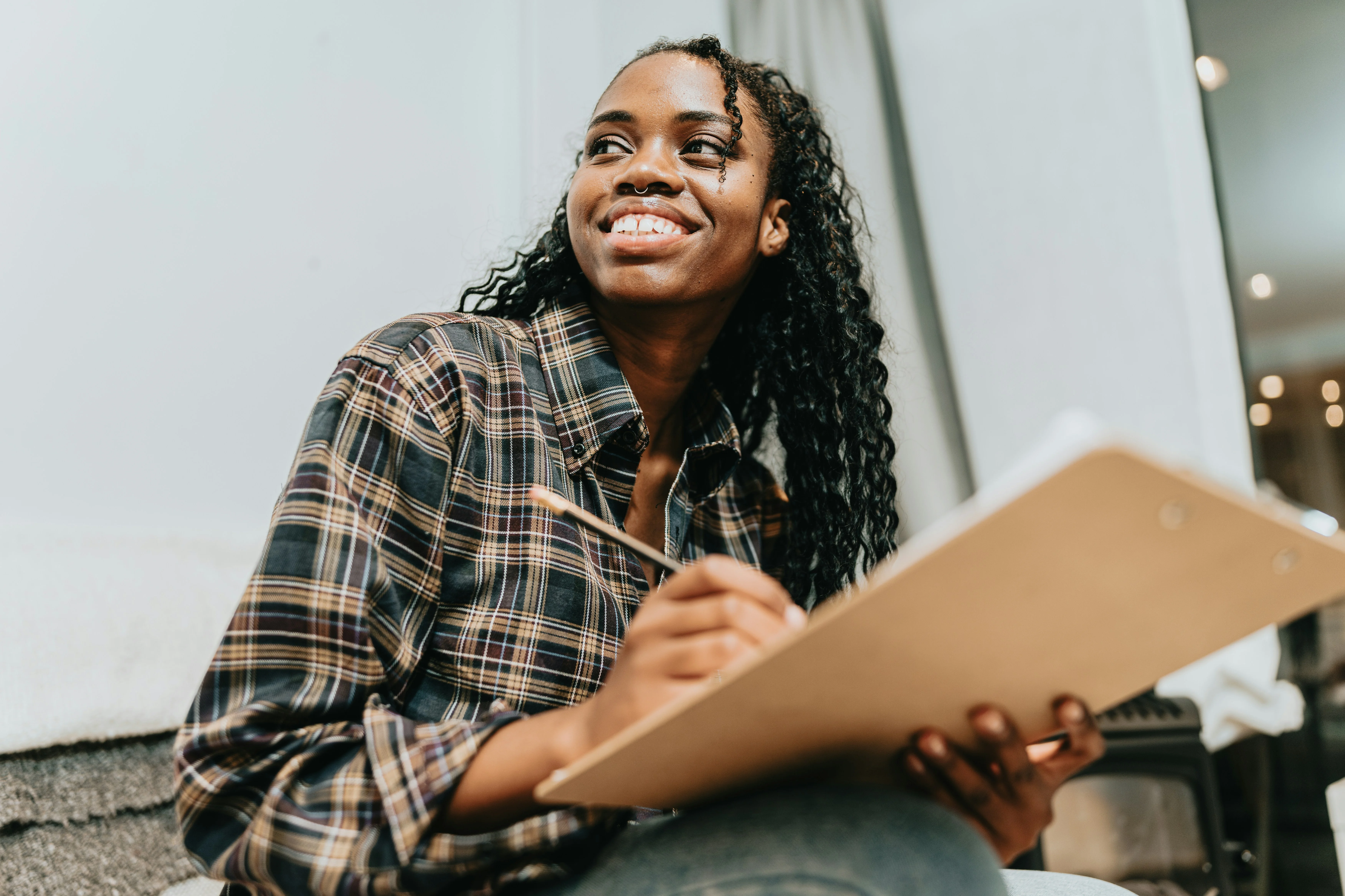 Smiling woman with long hair, wearing a plaid shirt, holds a sketchpad and pencil, looking upward thoughtfully.