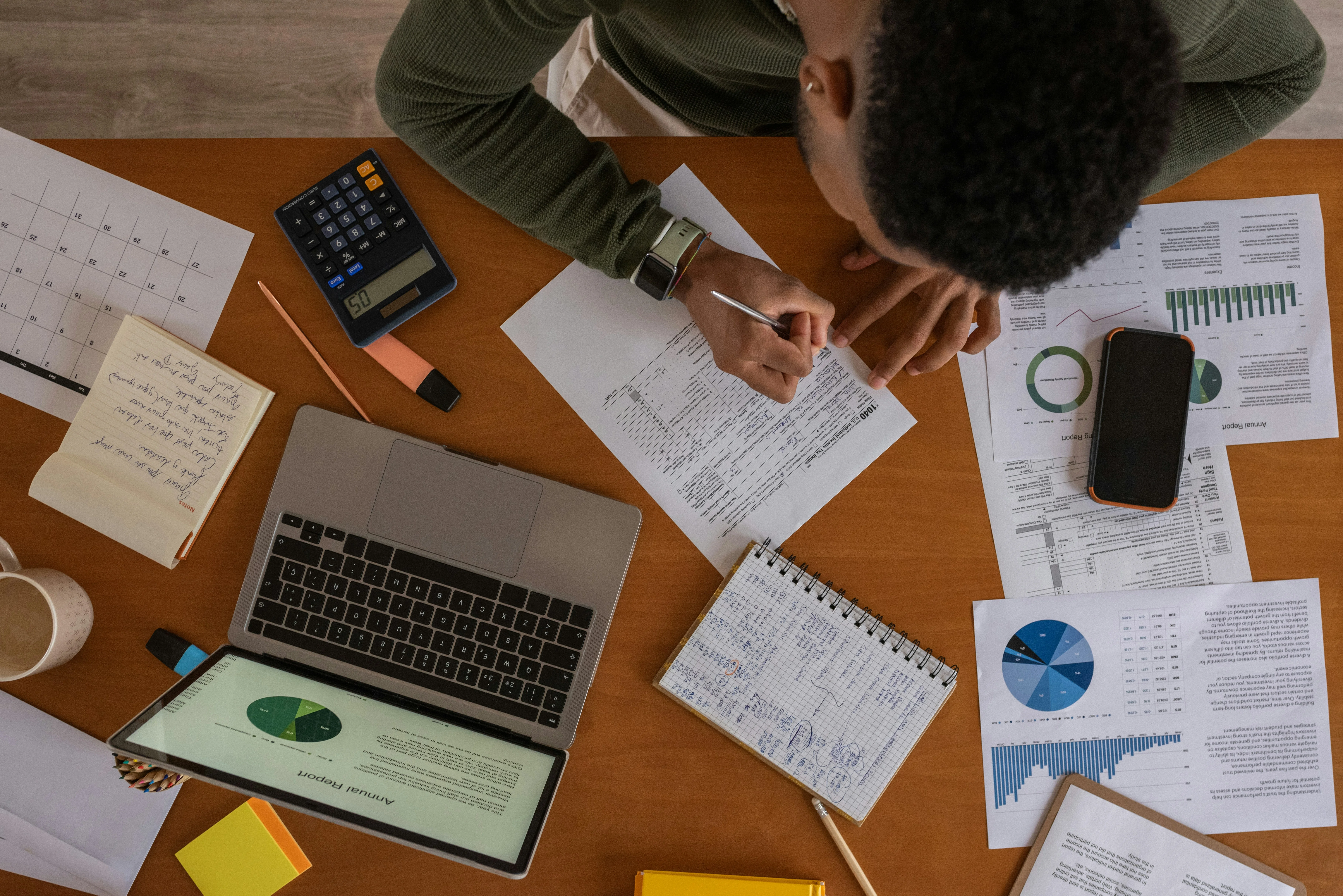 Person working at a desk with a laptop, calculator, charts, and papers. They are writing on a document amidst various office supplies.