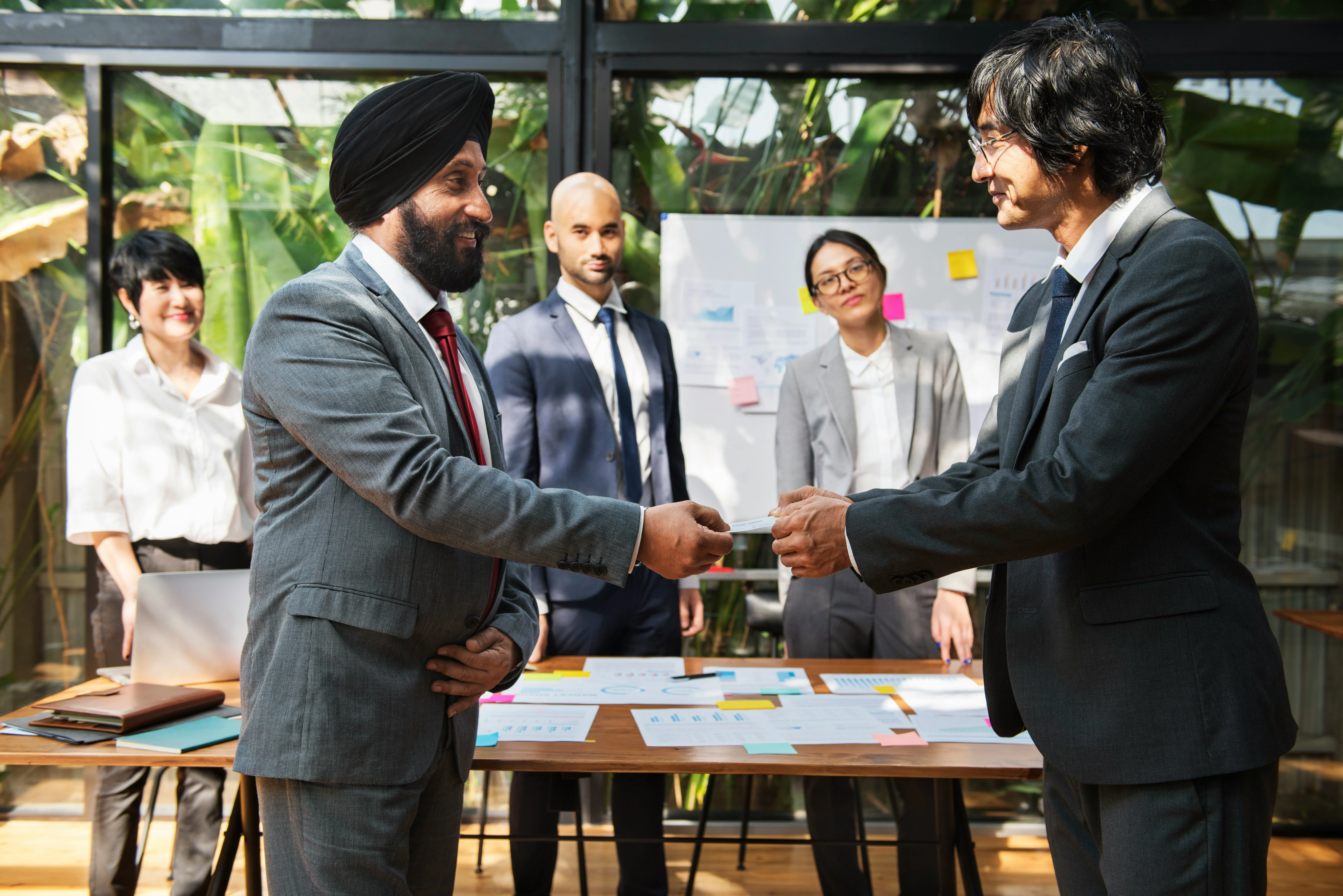 Two businessmen shaking hands in a meeting room, with three colleagues observing. A whiteboard and documents are visible in the background.