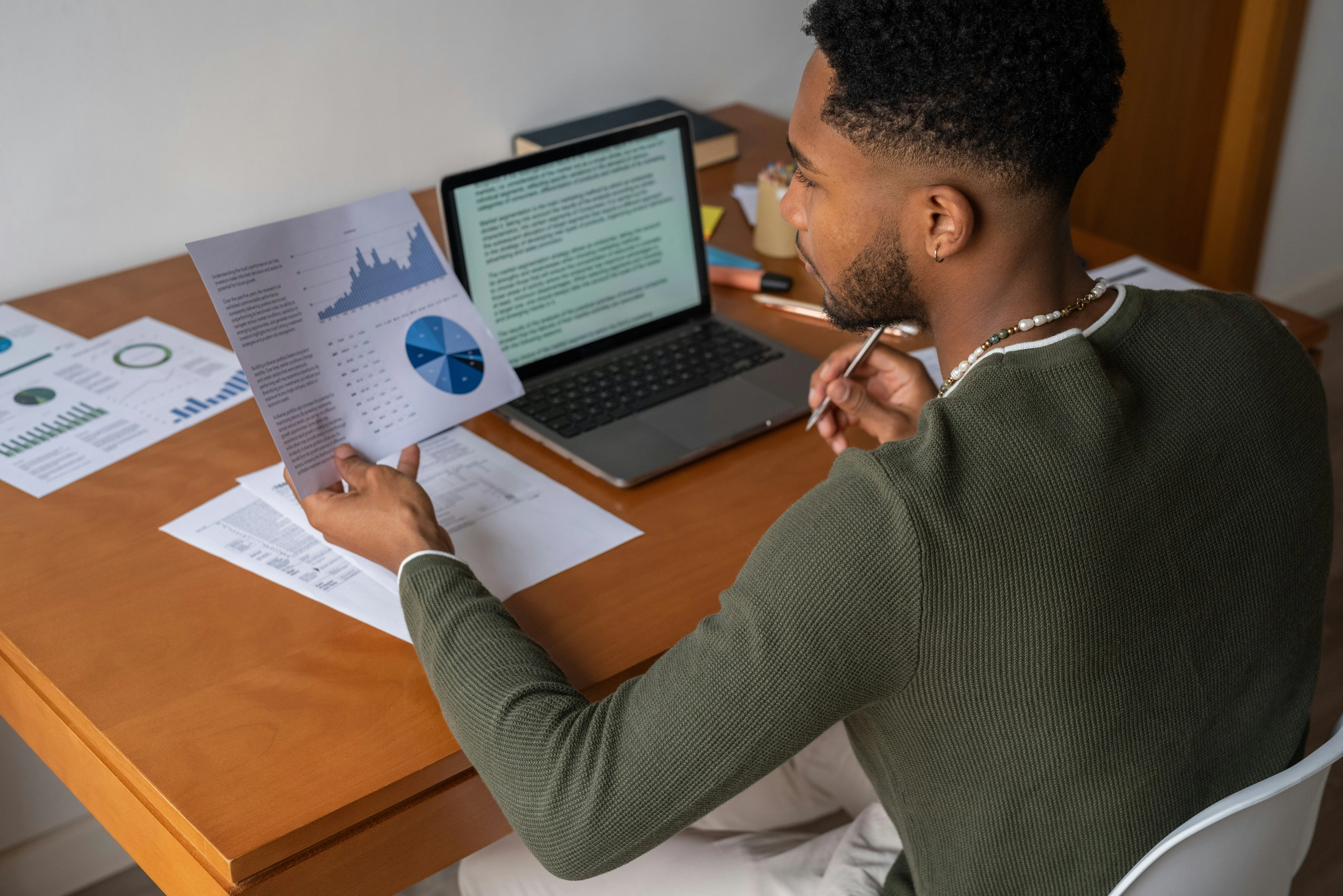 Man analyzing charts and graphs at a desk with a laptop, holding a paper with pie and bar charts, surrounded by documents.