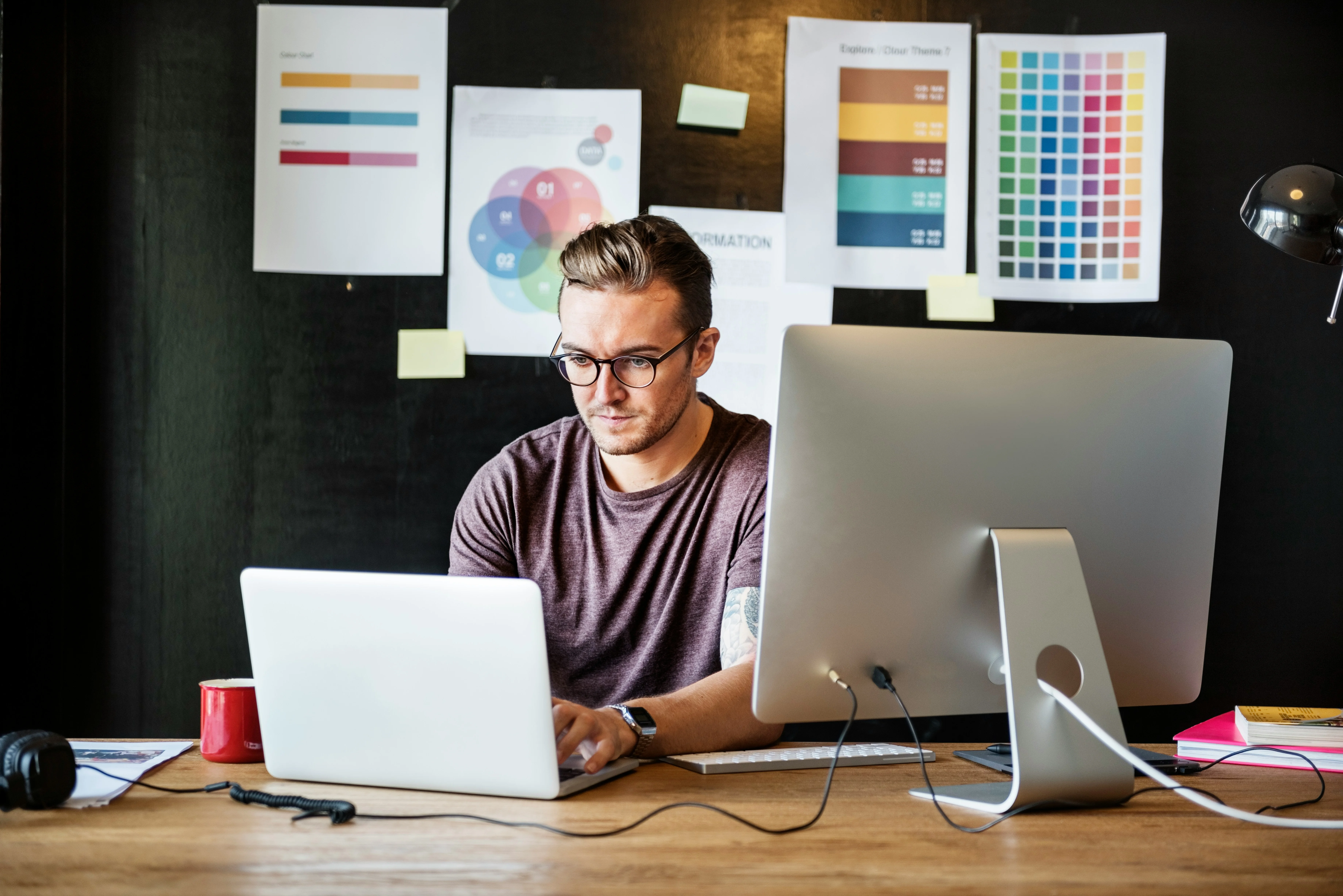 A person with glasses works at a desk with a laptop and monitor, surrounded by color charts and notes on a black wall.