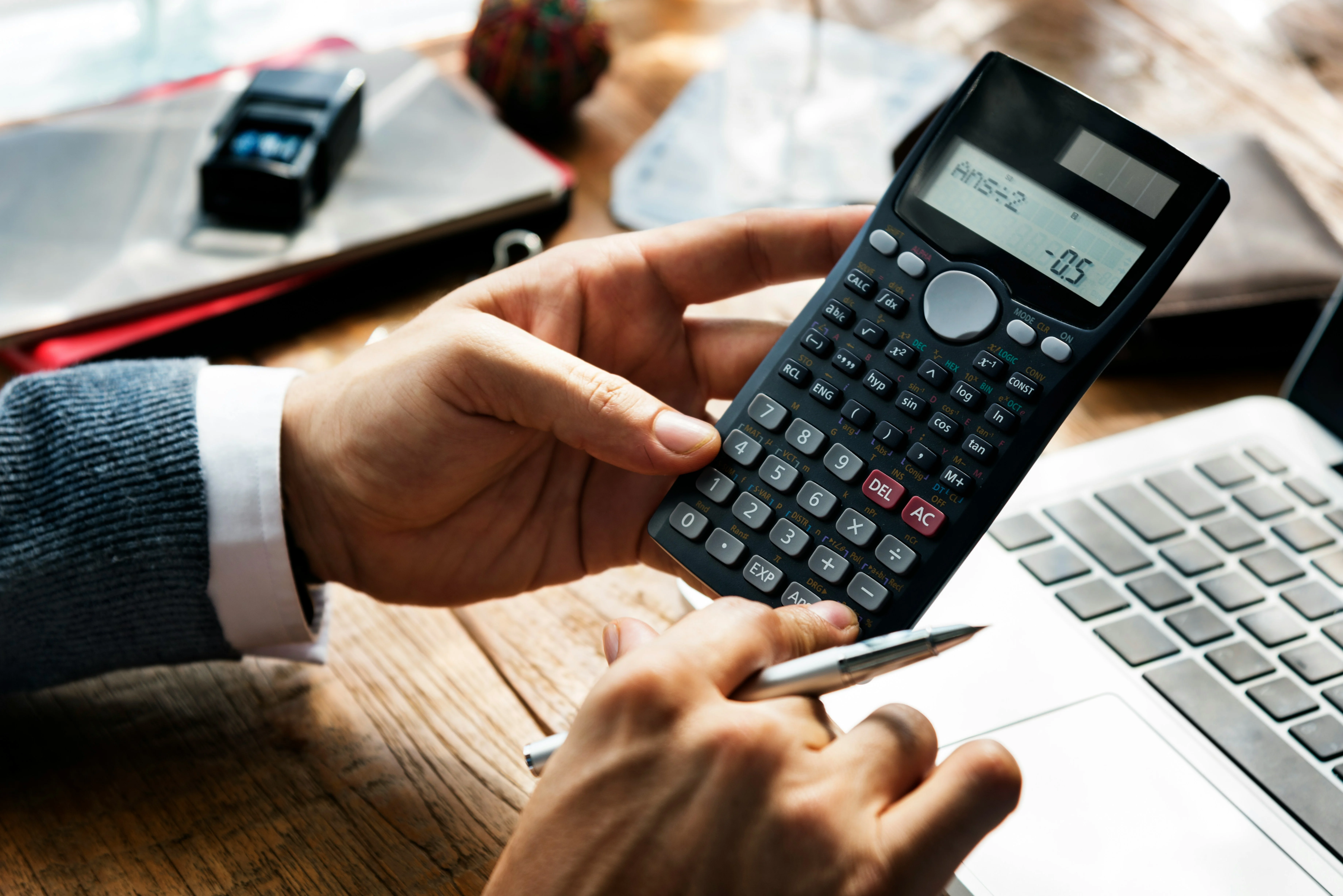 Person holding a scientific calculator, sitting at a desk with a laptop, pen, and documents.