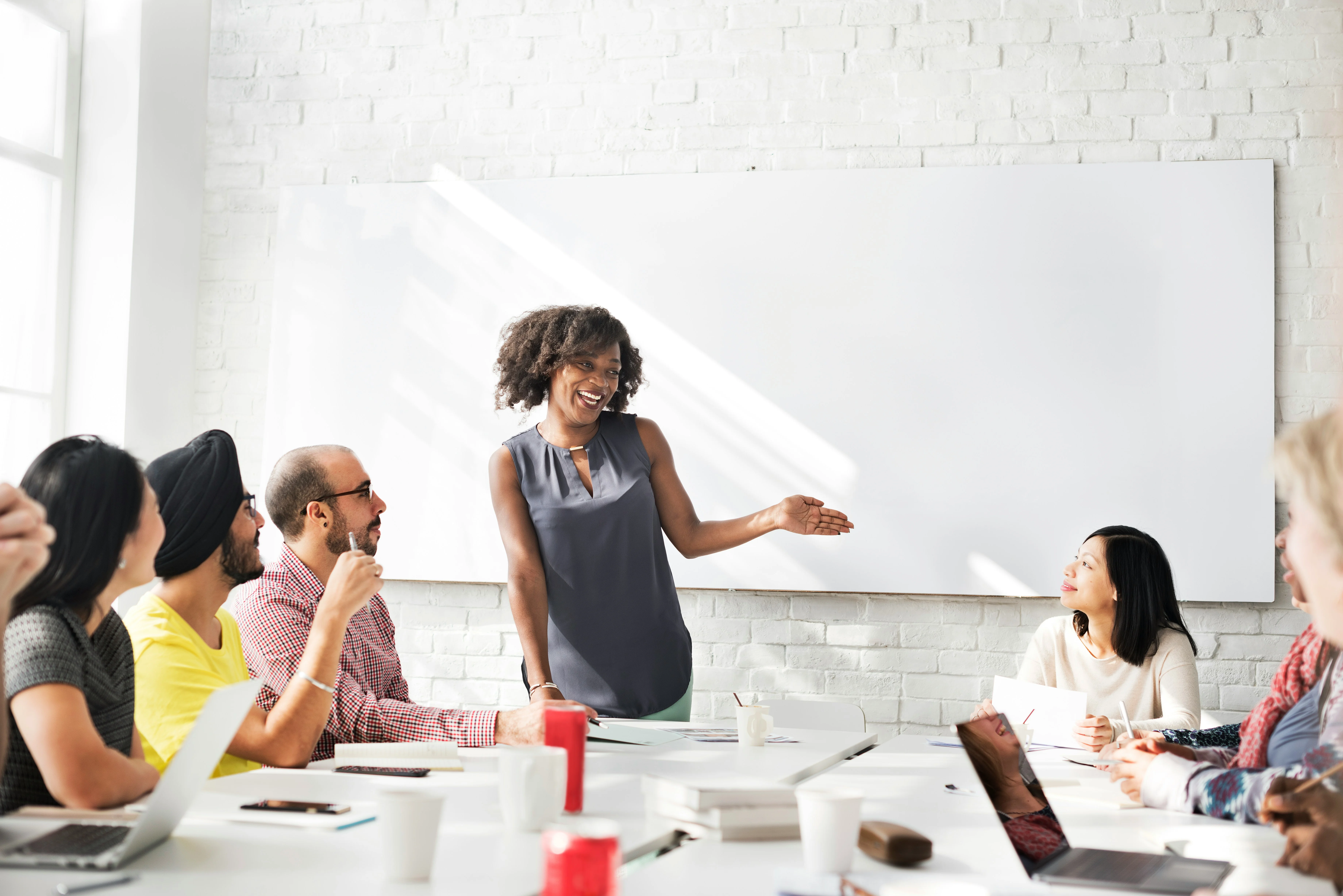 A woman stands and speaks to a group of people seated around a table in a bright meeting room with a whiteboard.