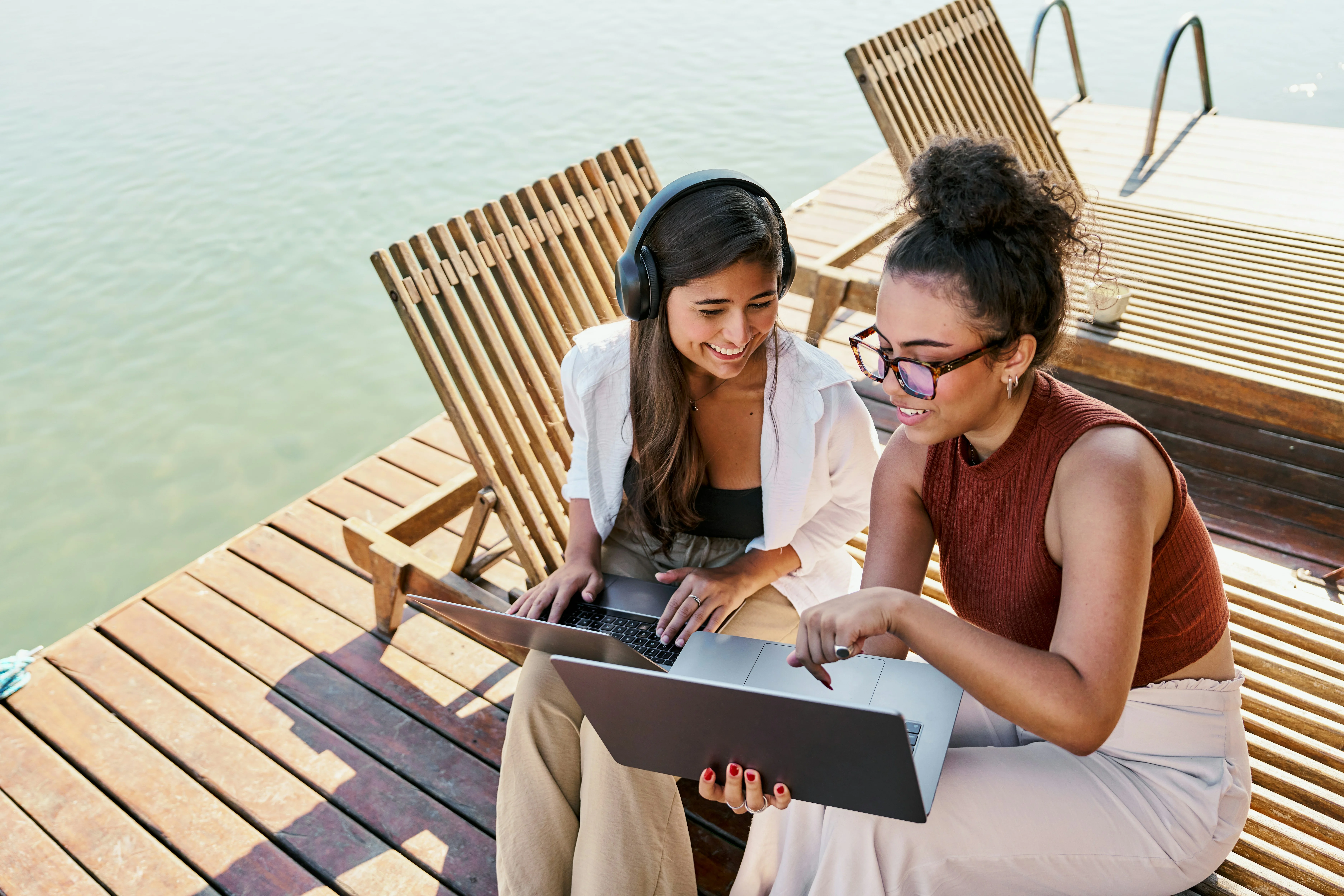 Two women enjoy working on a laptop together, sitting on wooden deck chairs by a lake. One is wearing headphones, both are smiling.
