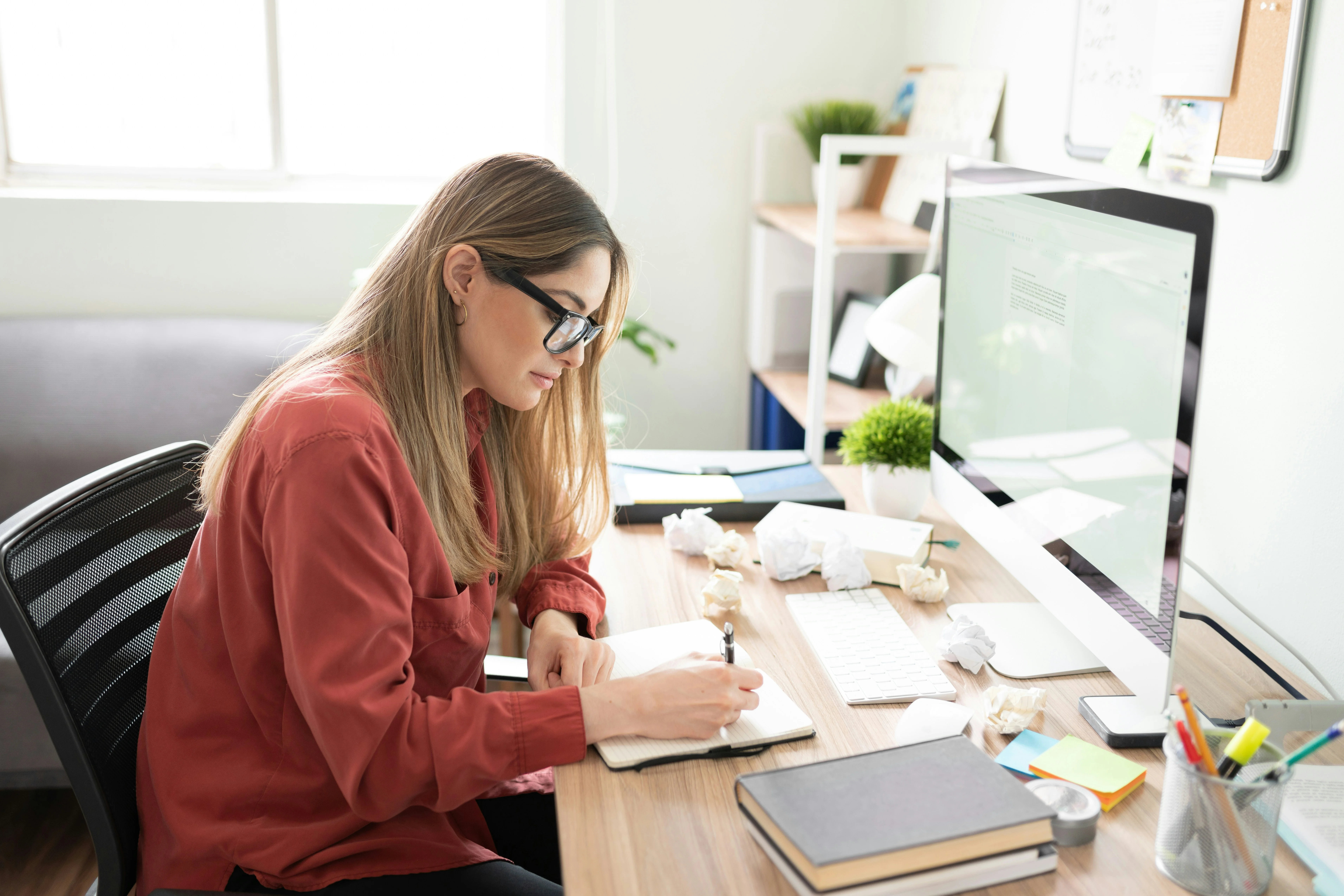Woman in glasses writing in a notebook at a cluttered desk with a computer, papers, and books in a bright room.