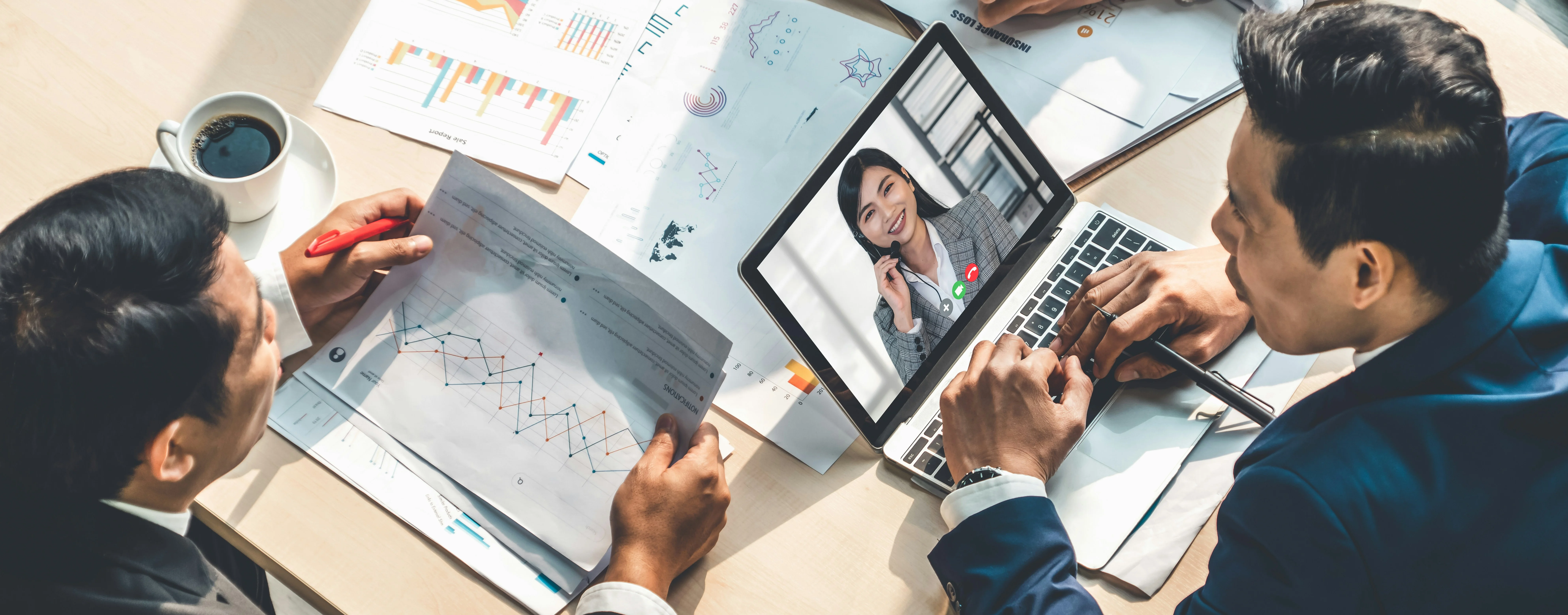 A virtual meeting with three people at a table, one on a laptop screen, discussing charts and graphs. A cup of coffee is nearby.