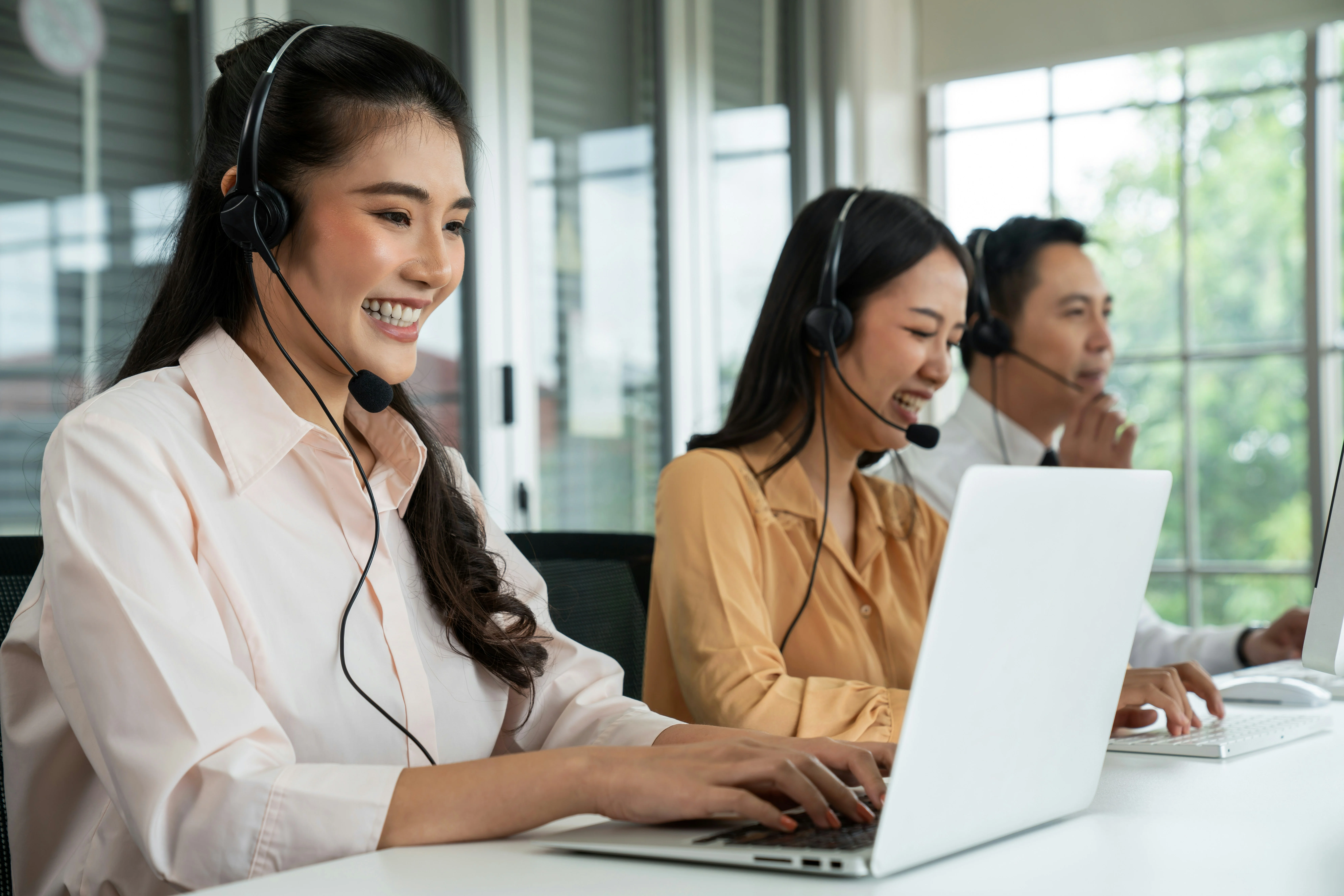 Three customer service representatives wearing headsets work on laptops in a bright office with large windows.