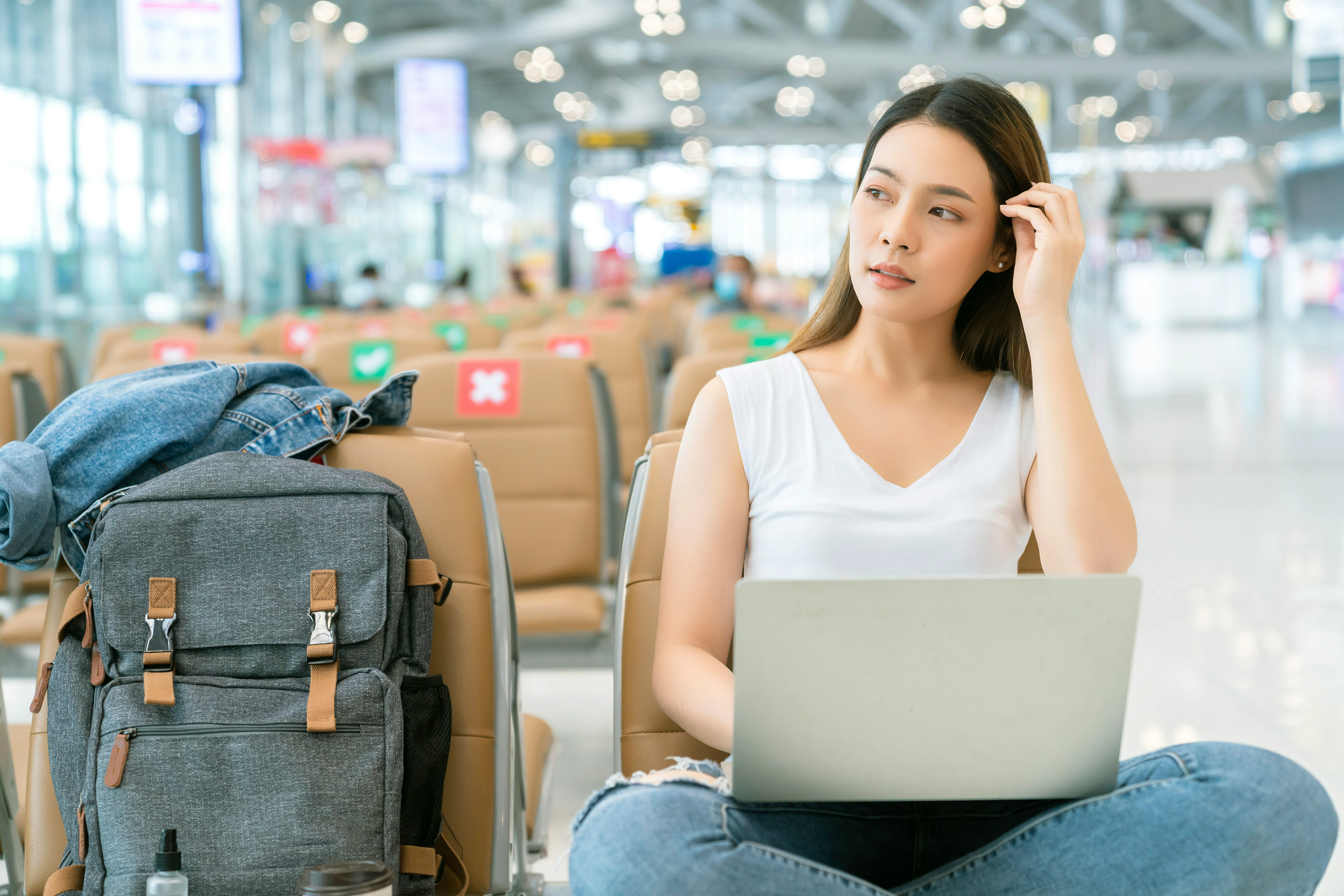 Woman in casual attire with a laptop sits in an airport terminal, surrounded by luggage and empty seats, looking thoughtfully to the side.