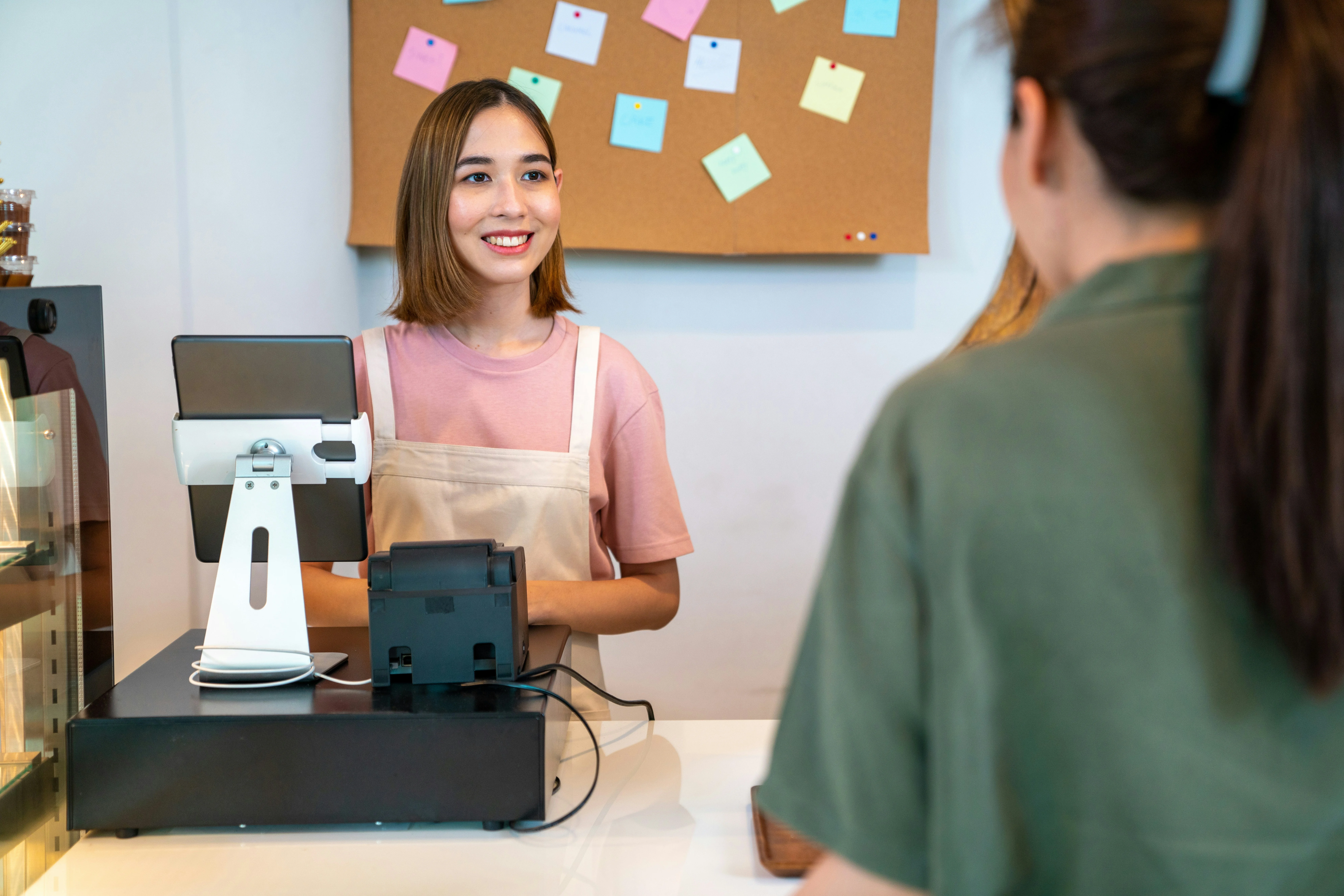 A smiling cashier in a pink shirt and apron assists a customer at a checkout counter, with colorful sticky notes on the wall behind.