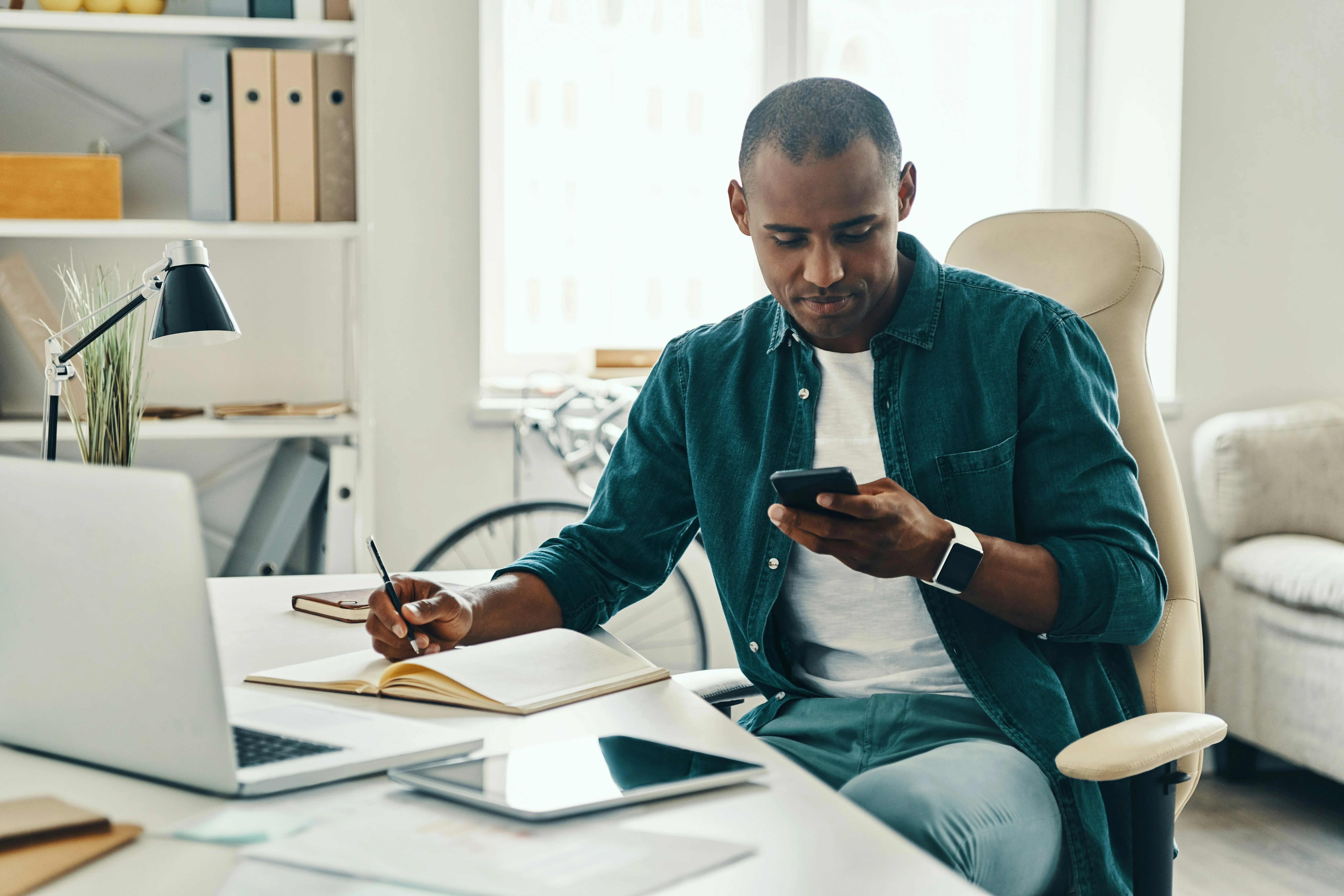 Man in a casual shirt sits at a desk, looking at his phone, with a notebook, laptop, and tablet nearby in a bright office space.