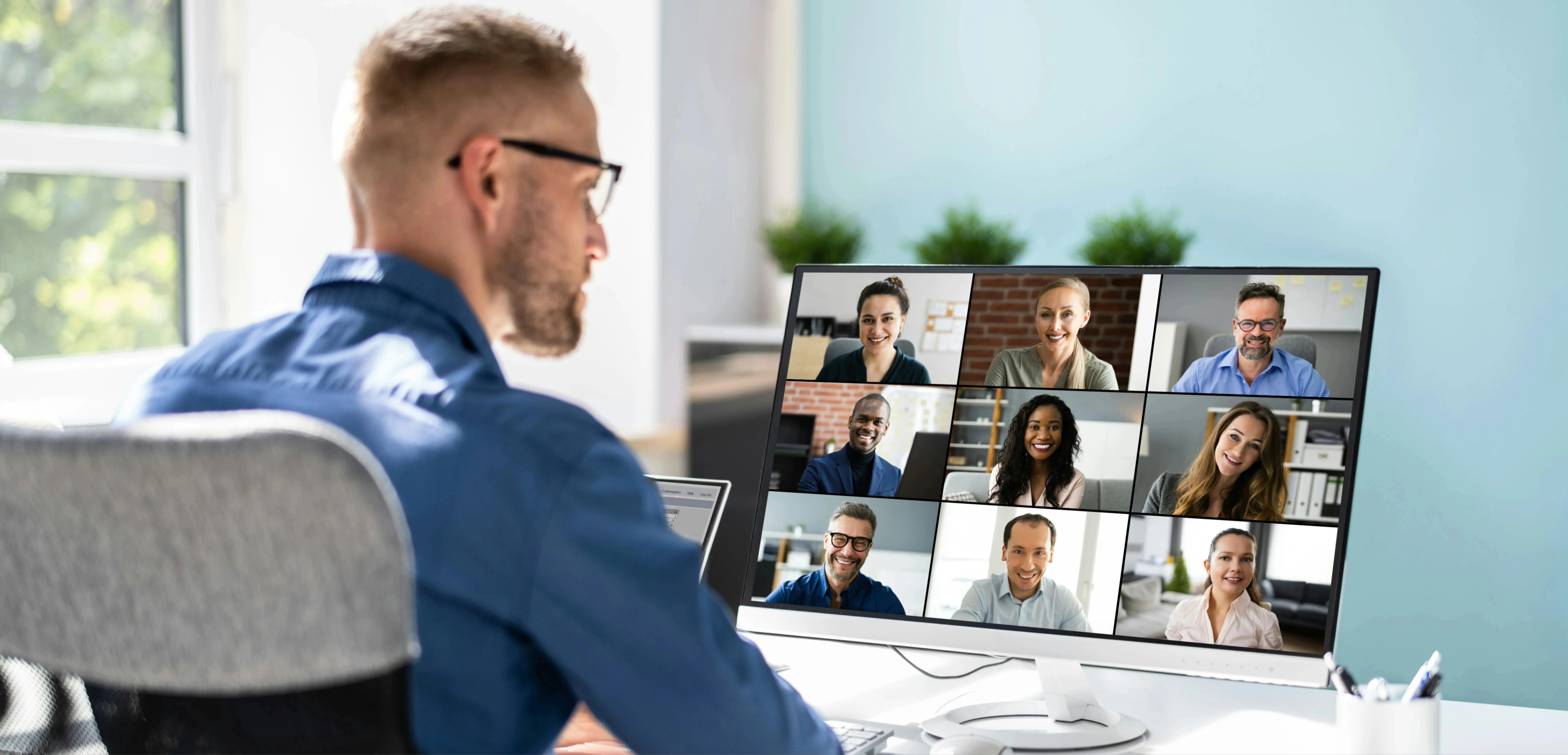 Man in blue shirt on a video call with nine people, displayed on a computer monitor, in a bright home office setting.