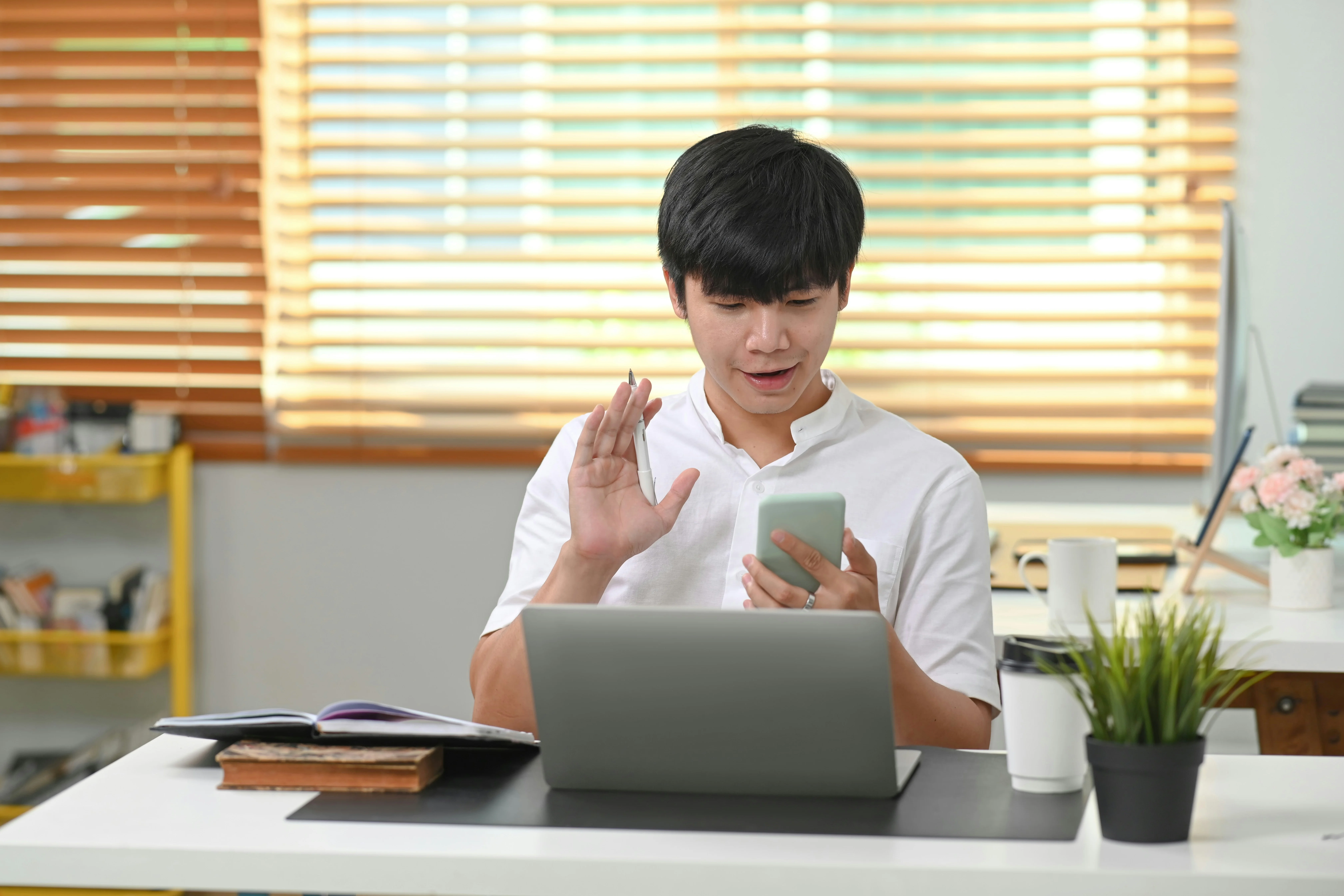 Man in white shirt video chatting on smartphone at a desk with a laptop, books, and a coffee cup, in a bright room with wooden blinds.