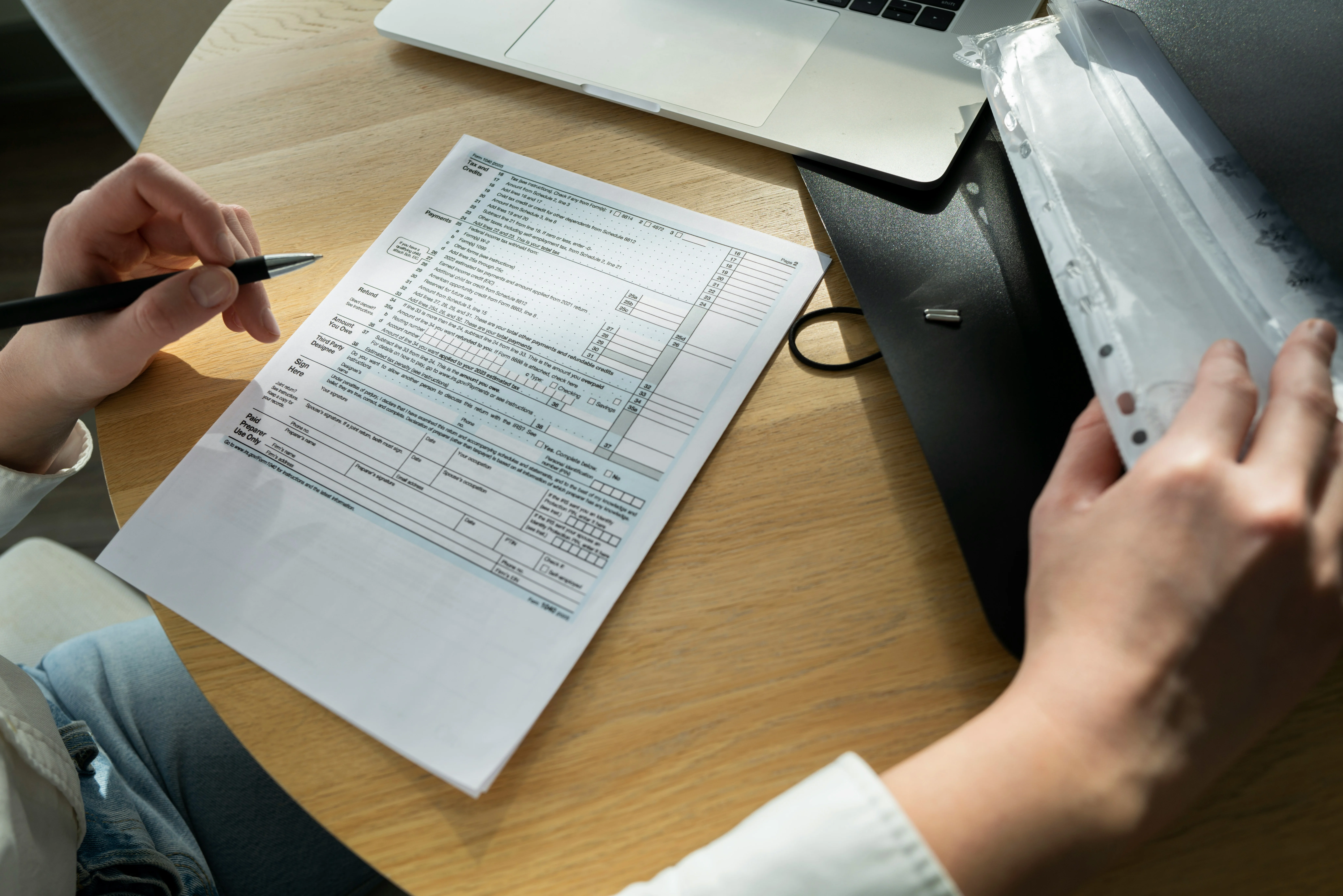 Person filling out a tax form at a wooden table, with a pen in hand, laptop open, and a folder nearby.