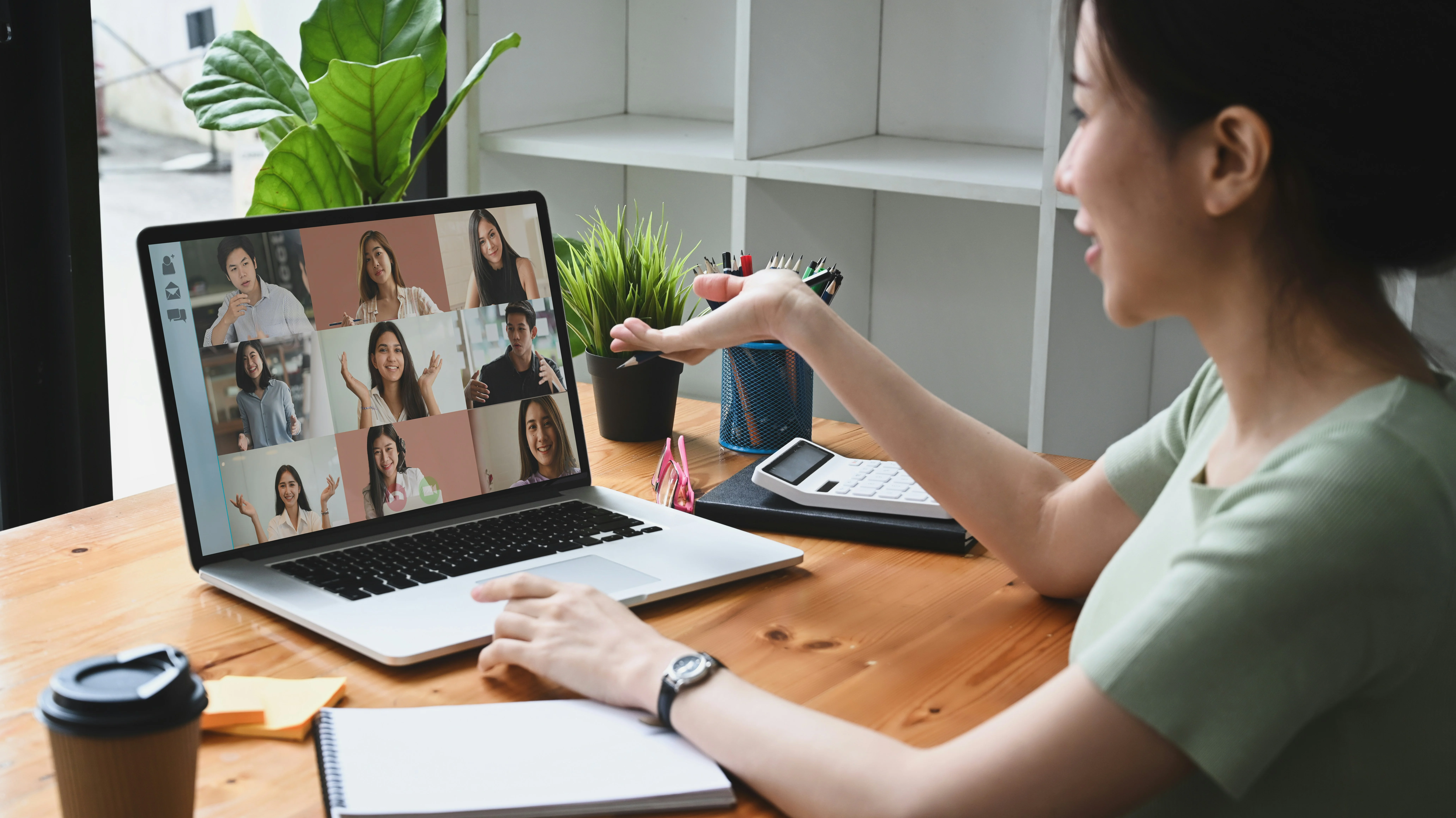 Woman gesturing during a video conference on a laptop at a desk with plants, a notebook, and a coffee cup. Participants are visible on the screen.