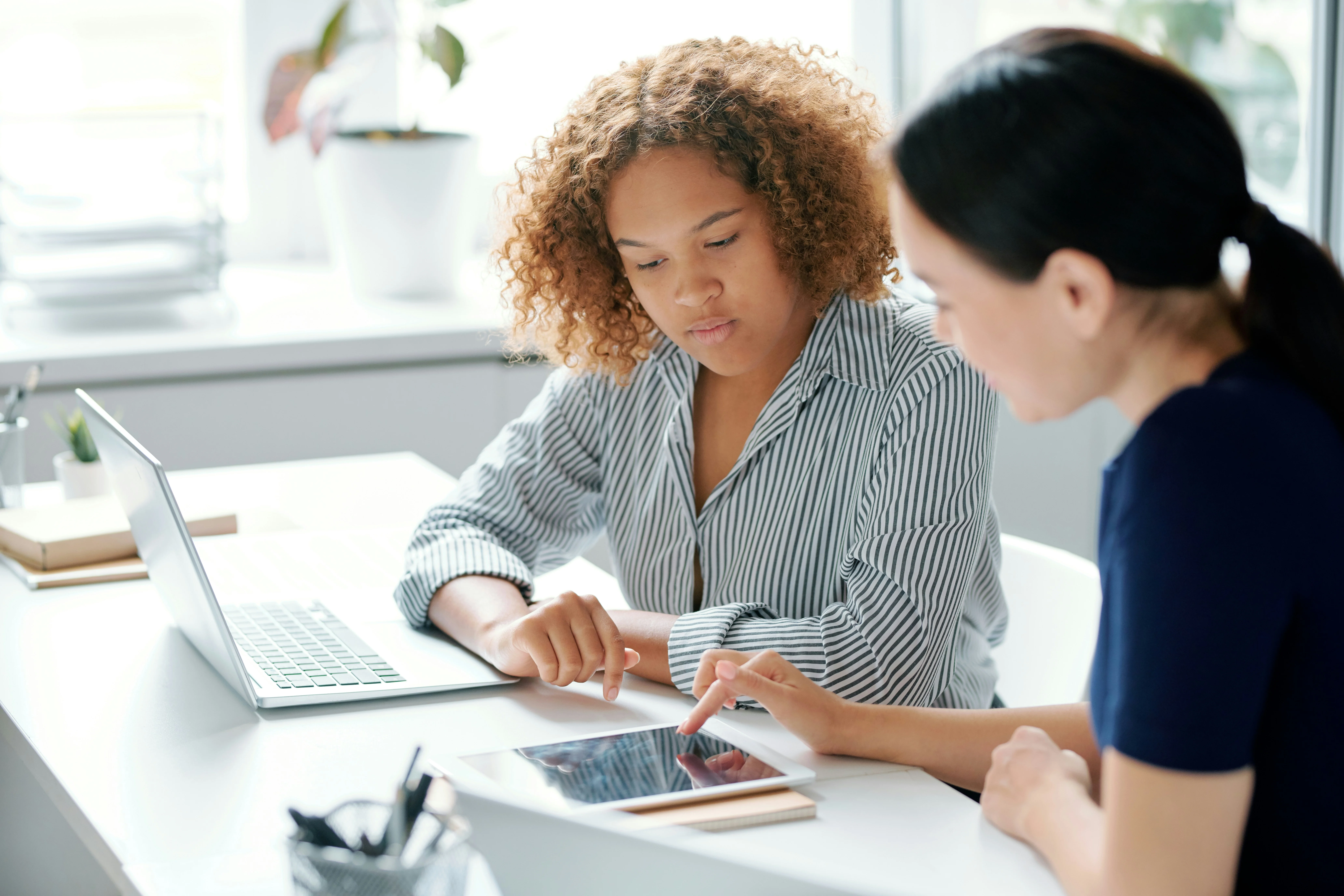 Two women sitting at a desk, one using a tablet and the other with a laptop. They appear focused and engaged in discussion.