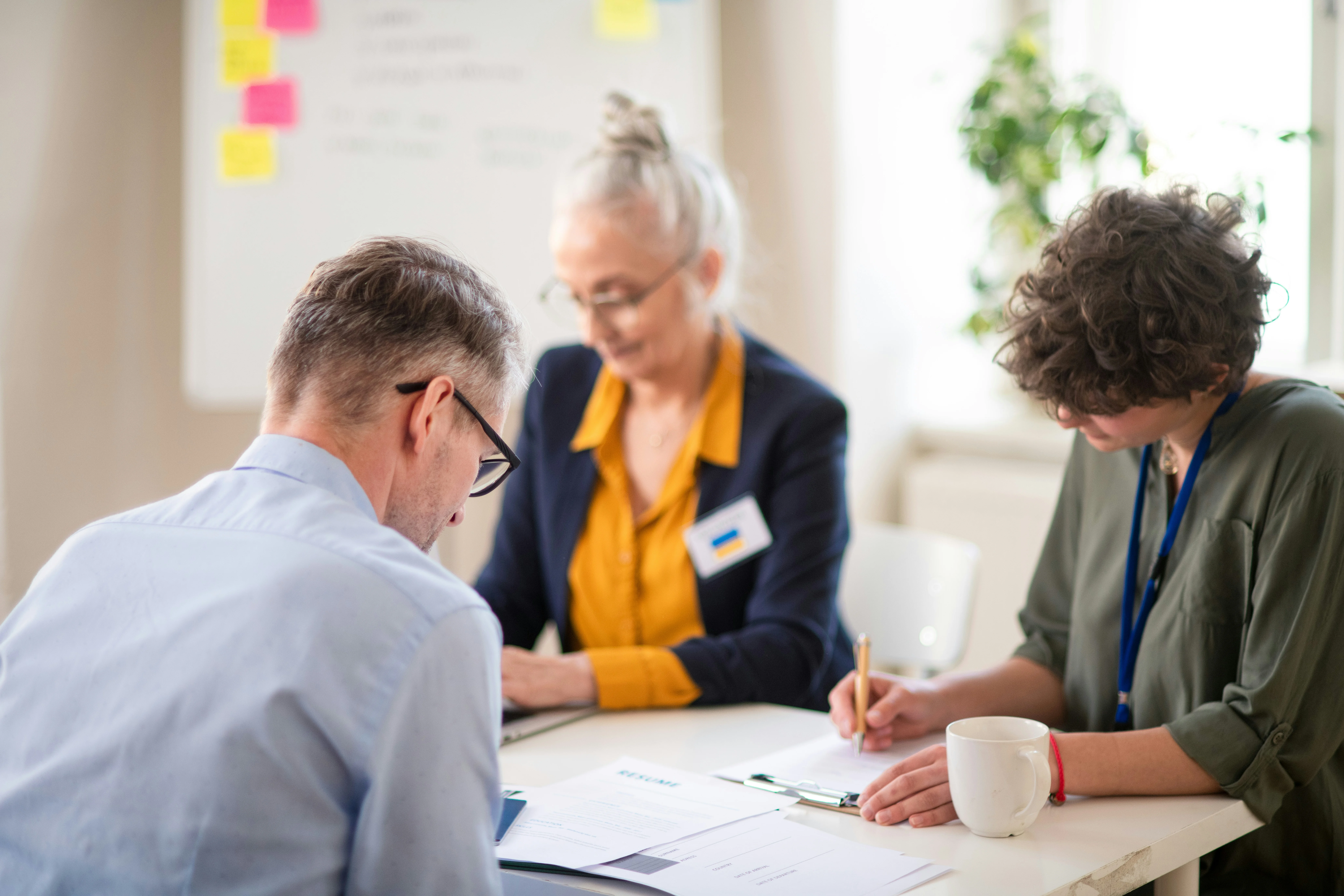 Three people sit at a table, focused on writing notes. A whiteboard with sticky notes is in the background.
