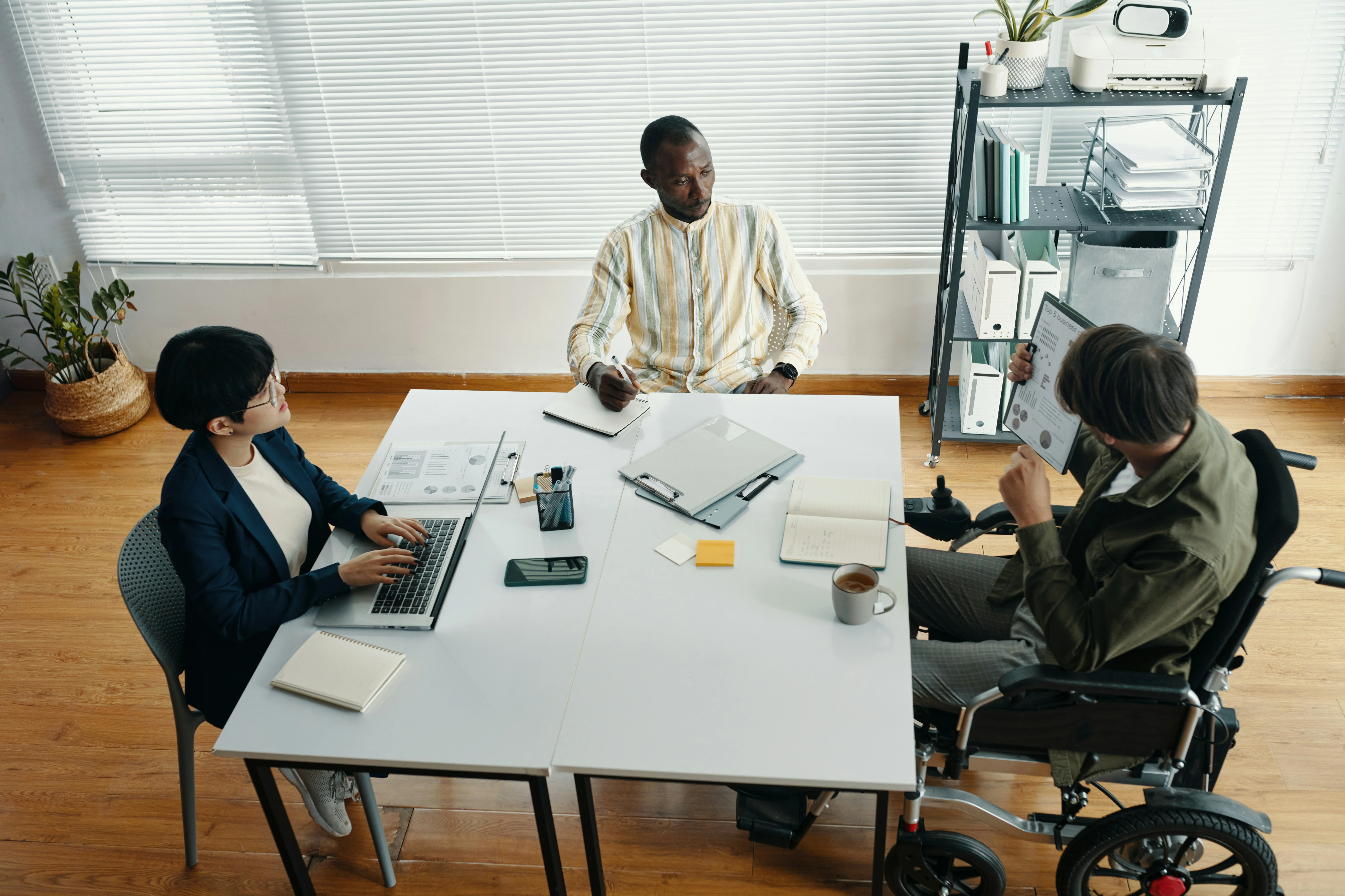 Three people in a meeting room: one using a laptop, another taking notes, and one in a wheelchair showing a document.
