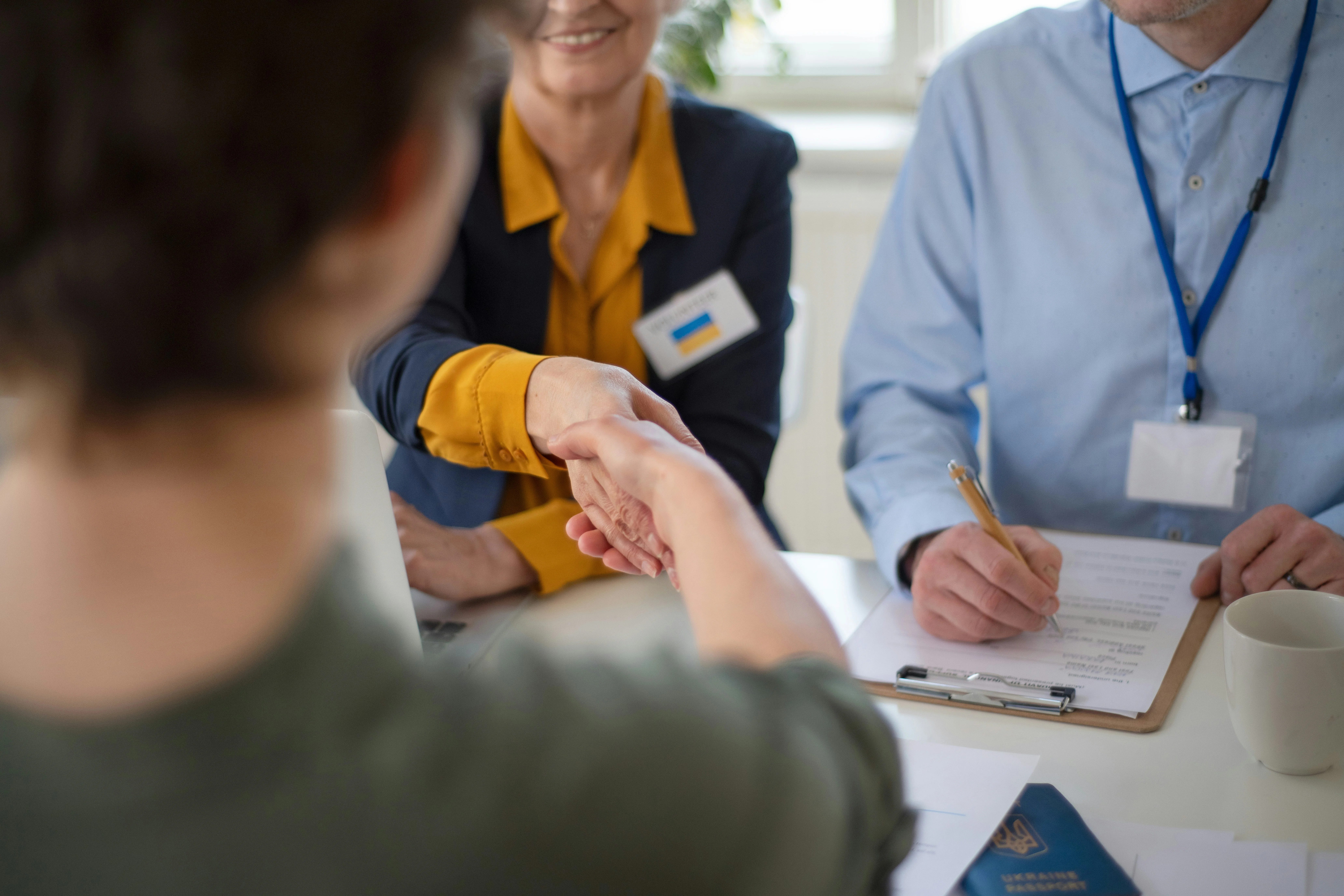 Person shaking hands with another person across a table, while a third person takes notes on a clipboard.