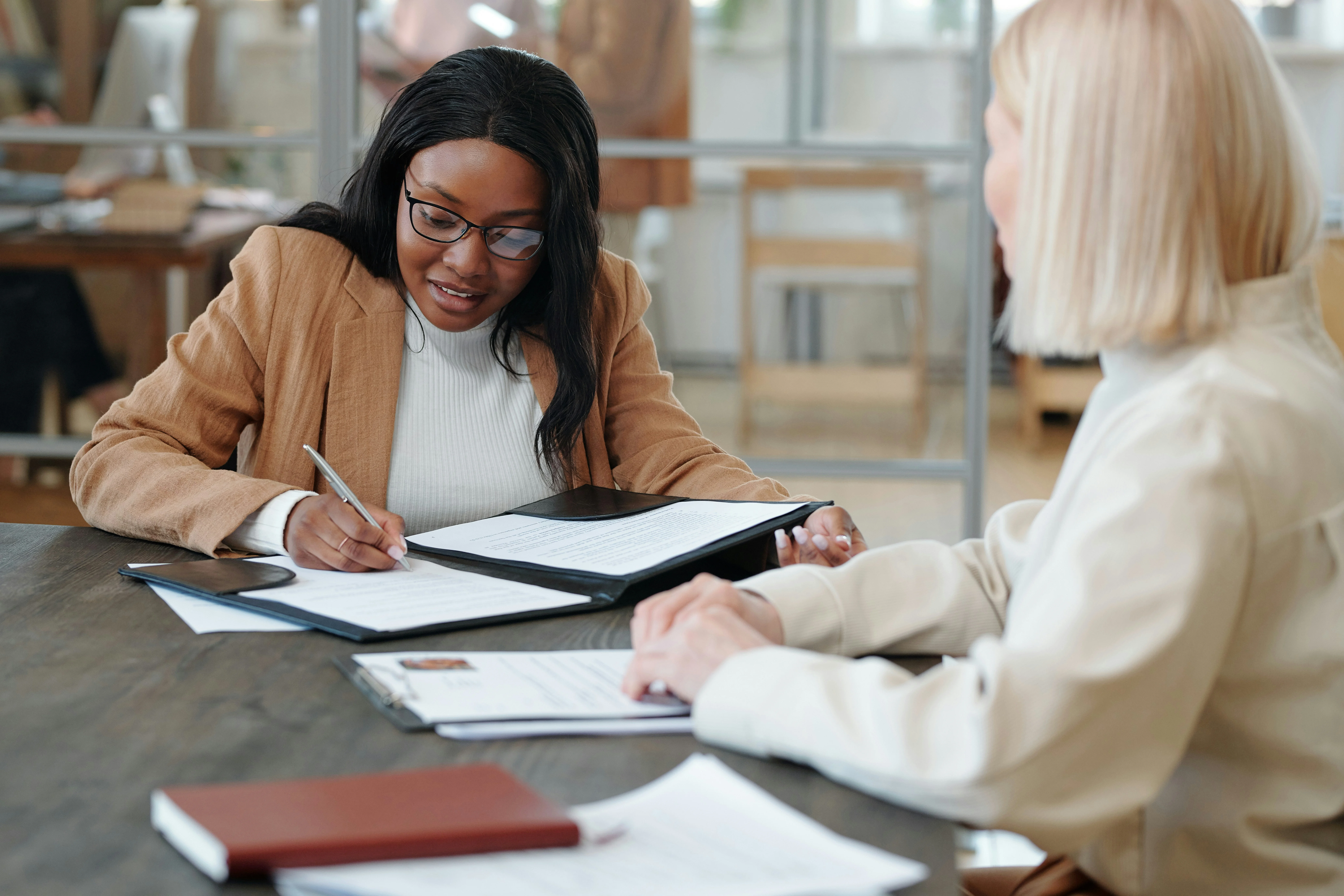 Two women seated at a table in an office, discussing and reviewing documents. One is writing, while the other listens attentively.