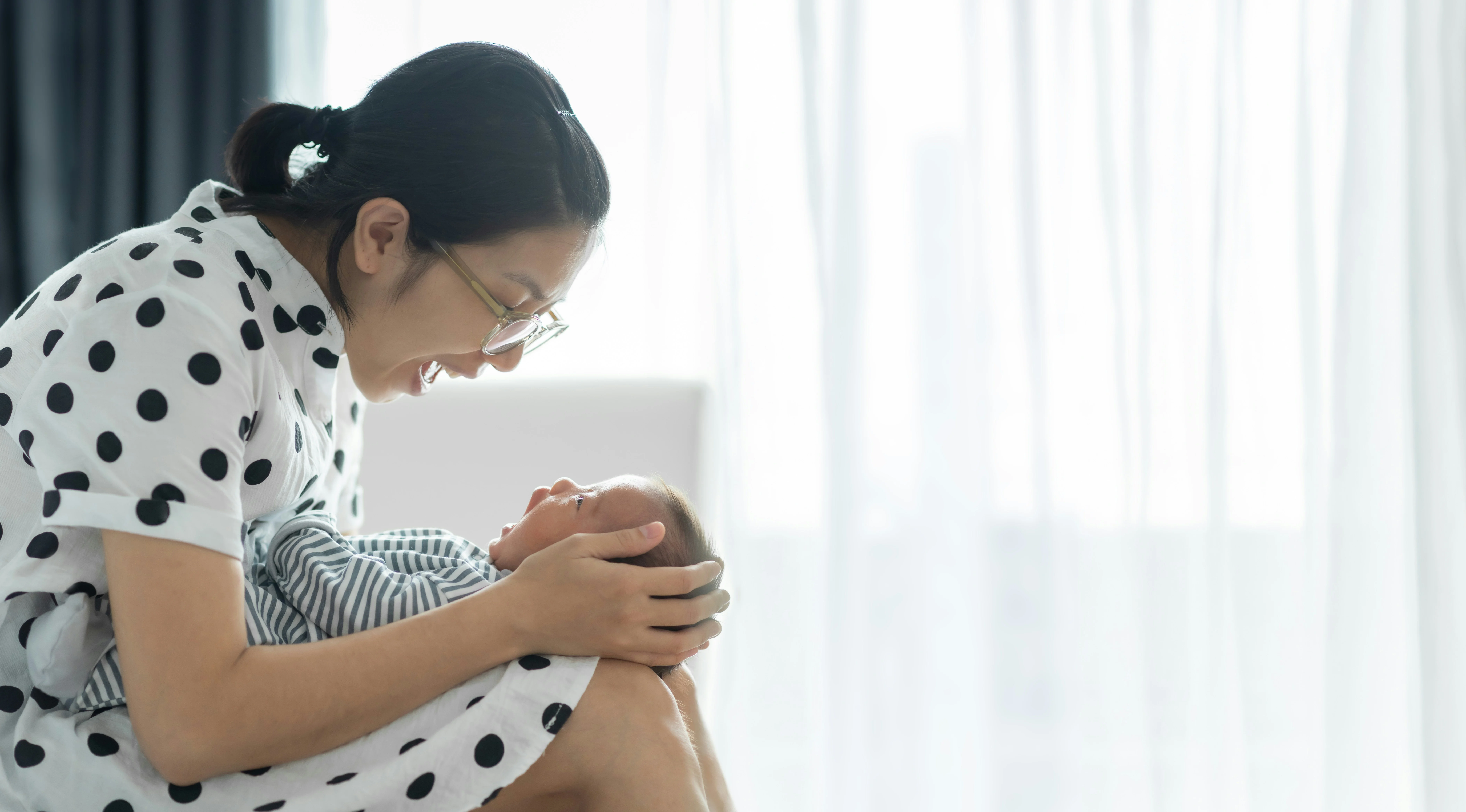 A woman in a polka dot dress smiles at a baby wrapped in a striped blanket, sitting in a softly lit room.
