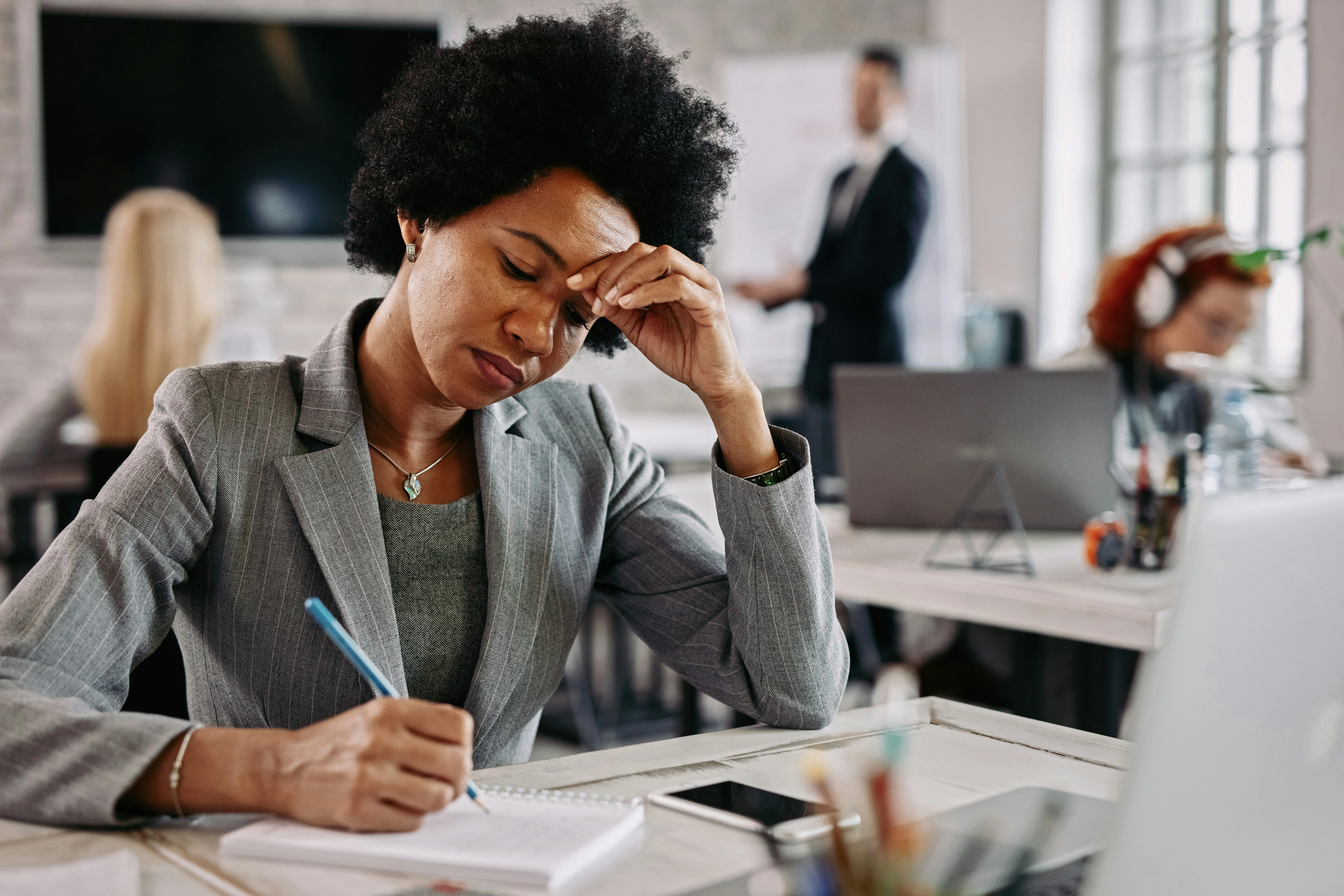 A woman in a suit looks stressed, holding her forehead while writing in a notebook at an office desk. Colleagues work in the background.
