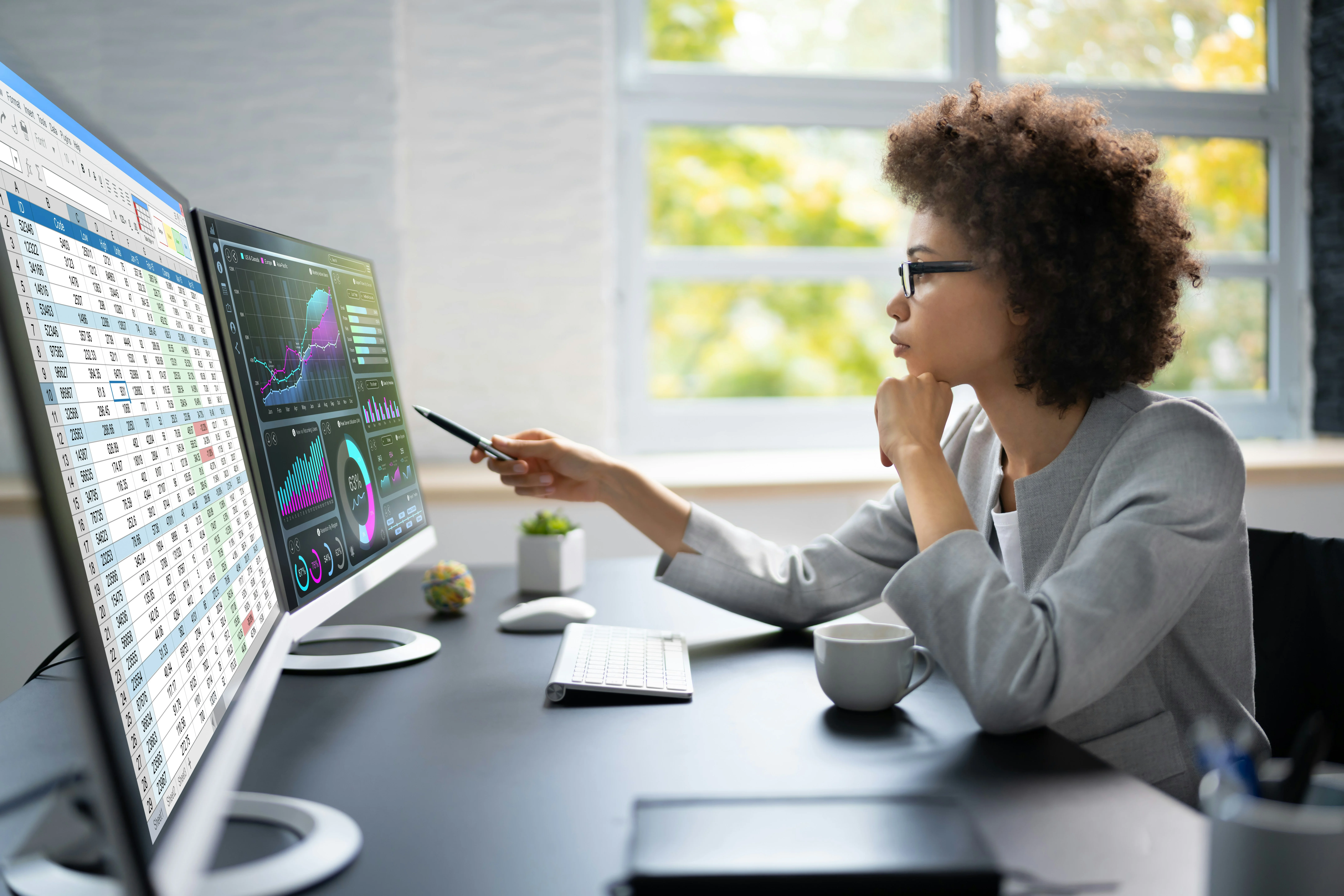 A person in an office analyzes data on dual monitors, displaying spreadsheets and graphs, while holding a pen and drinking coffee.