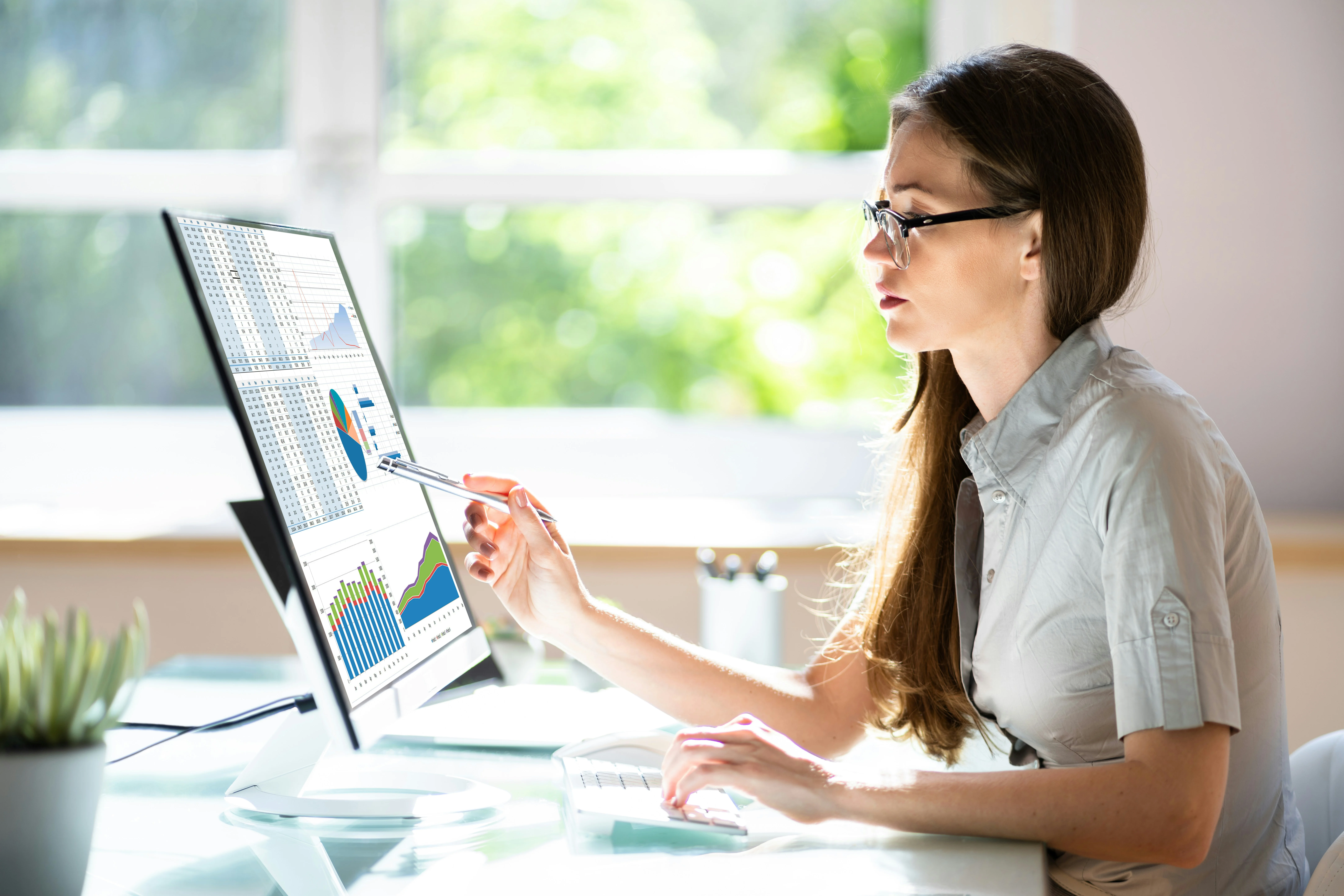 A woman in glasses uses a stylus to point at graphs on a computer screen in a bright office.