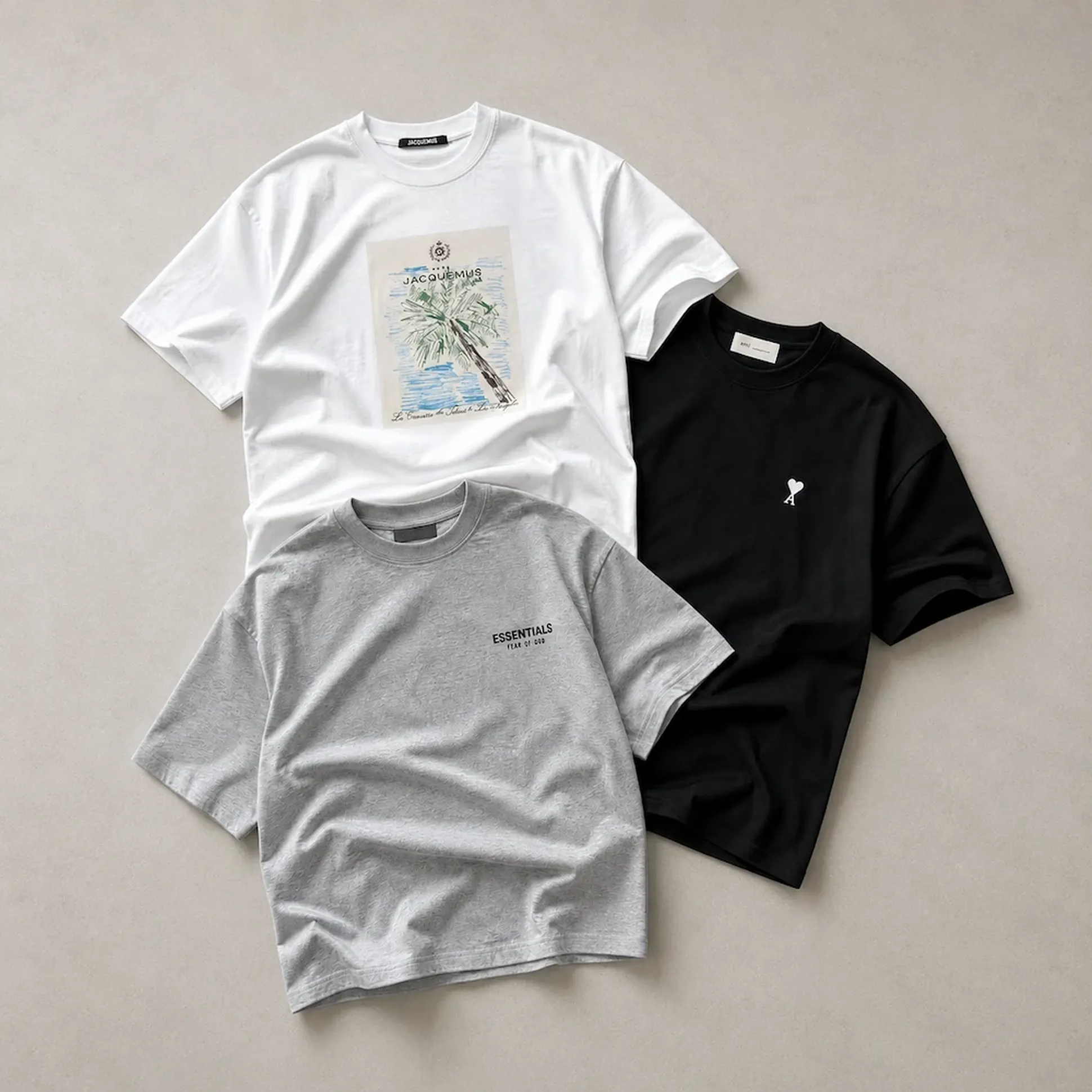 Three T-shirts on a flat surface: white with palm tree print, gray with "ESSENTIALS" text, and plain black with small logo.